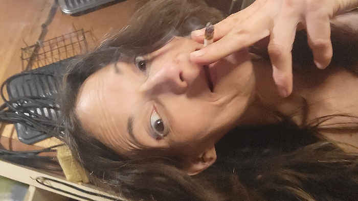 A woman with long, dark, wavy hair holds a lit cigarette or joint to her lips. She looks directly at the camera with wide eyes. She is light-skinned with visible pores and wrinkles. Smoke rises from the end of the cigarette. In the background, there is a black grill and a wire rack on a wooden surface. The image is slightly tilted.