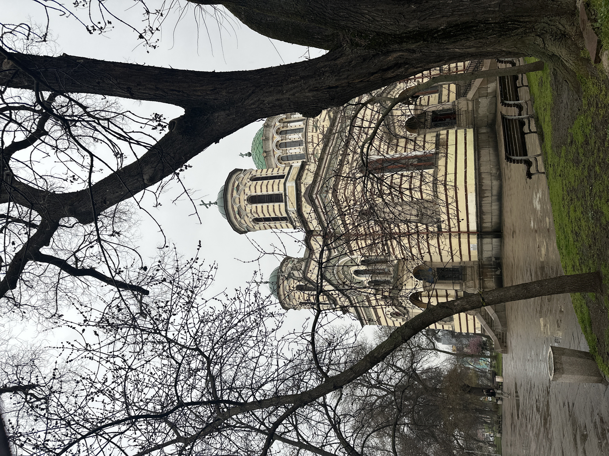 The image depicts the Alexander Nevsky Cathedral in Sofia, Bulgaria, partially obscured by bare tree branches in the foreground. The cathedral's ornate facade and domes are visible. To the right, a paved area is lined with benches and some greenery. The sky is overcast. No people are clearly visible.