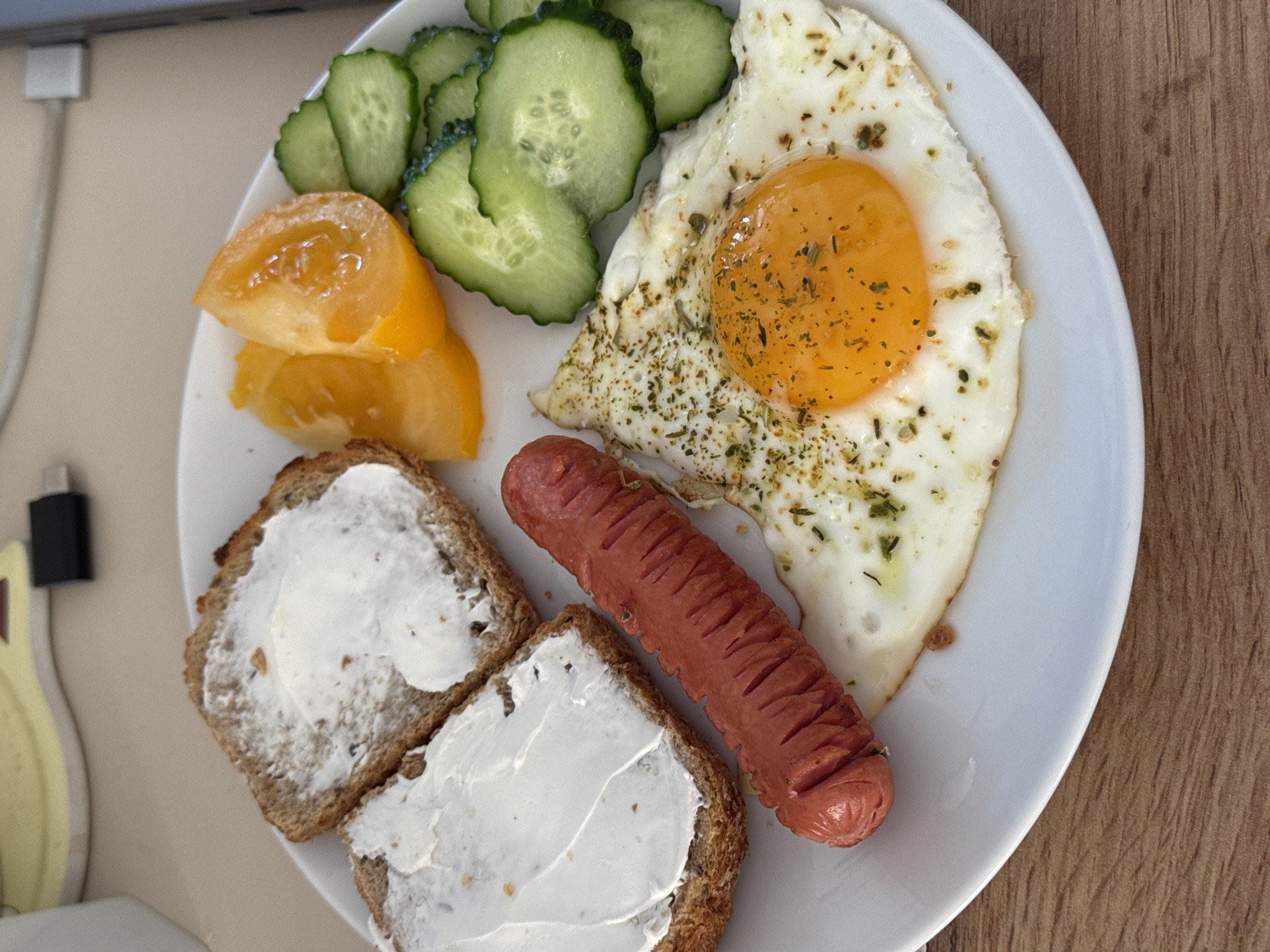 A plate of food is centered on a wooden surface. The plate contains two slices of whole wheat bread, each spread with a thick layer of white cream cheese. Next to the bread is a single, long, reddish-brown sausage, slit lengthwise. A fried egg, with a bright yellow yolk and white, is placed beside the sausage, seasoned with herbs. To the right of the egg are several slices of cucumber and wedges of yellow tomato. The background shows a blurry off-white surface with a black electronic device visible. The lighting suggests an indoor setting.