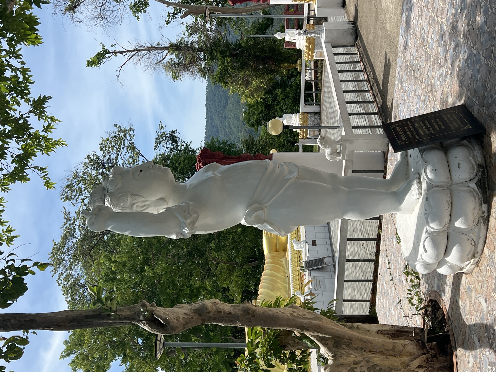 In Baan Thong Krut, Thailand, a white stone statue of a cherubic figure, possibly a deity or child, stands prominently on a lotus-shaped pedestal. The statue is depicted in profile, facing right, with its right arm raised, holding an unseen object or in a gesture of offering. The figure wears a simple cloth around its waist. Behind the statue, a golden reclining Buddha statue is visible, lying on a raised platform. The scene is set outdoors, with lush green trees and a clear blue sky with scattered clouds forming the backdrop. A tiled pathway surrounds the statues, and a black informational plaque with ornate gold lettering stands in the foreground, to the right of the cherubic statue. The overall atmosphere suggests a religious or spiritual site, likely a temple or sanctuary.