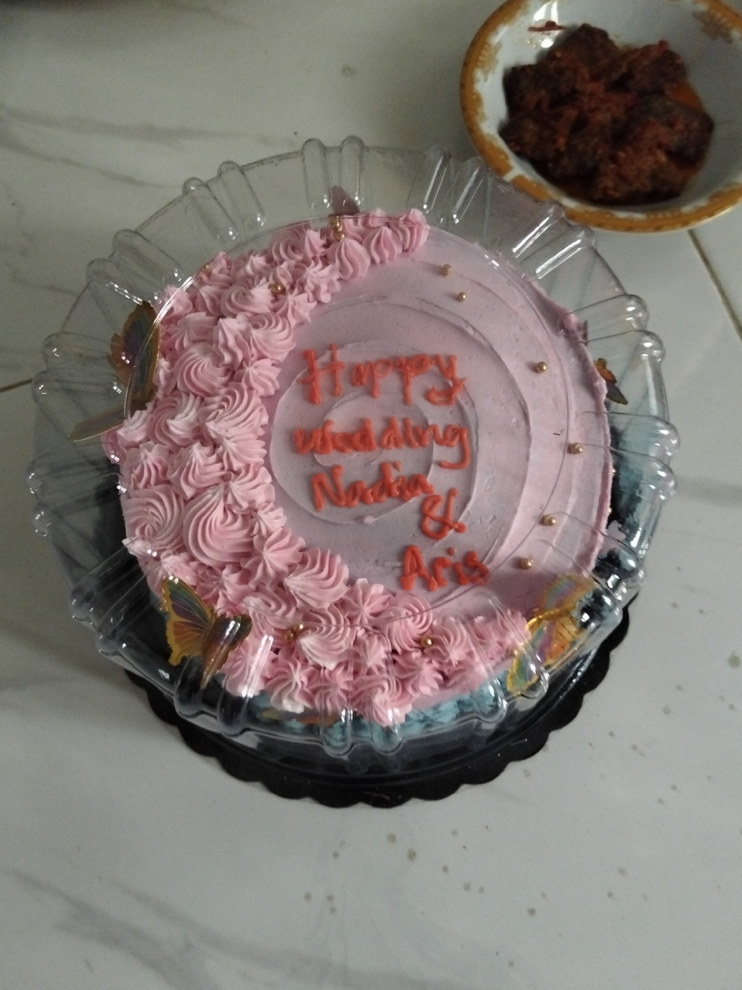 A pink frosted cake sits in a clear plastic container on a marble-patterned surface. The cake is decorated with piped pink frosting around the edge and has "Happy Wedding Nadia & Aris" written in orange icing in the center. Several edible gold balls are scattered on the cake. Two decorative butterflies, one orange and one iridescent, are attached to the side of the cake. A small bowl of what appears to be food is visible in the upper right corner of the image. The scene is indoors, suggesting it might be a home or a bakery in Cottrueng, Indonesia.