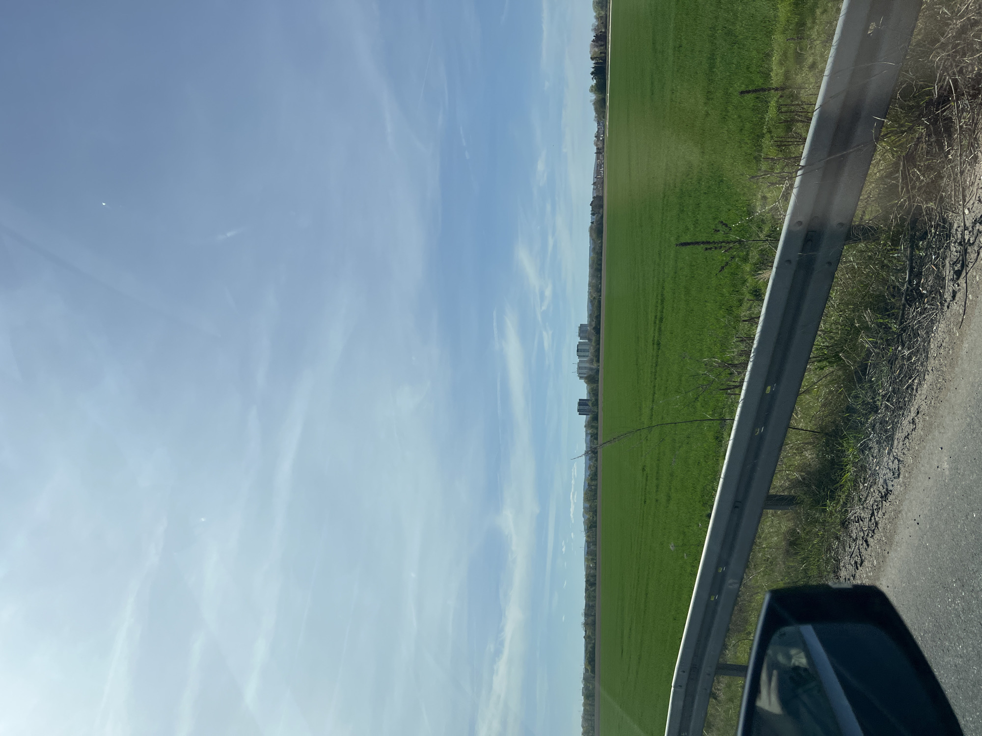 The image depicts a view from a moving vehicle, likely a car, traveling on a road or highway in what appears to be a rural or semi-rural area. The sky is light blue with scattered wispy clouds. To the right of the frame is a green field, possibly farmland. A guardrail runs along the edge of the road, separating it from the field. The road surface is asphalt. In the foreground, a portion of the car's side mirror is visible. In the distance, there are buildings visible. Based on the information provided, the location is likely somewhere near Eschweiler, Germany.