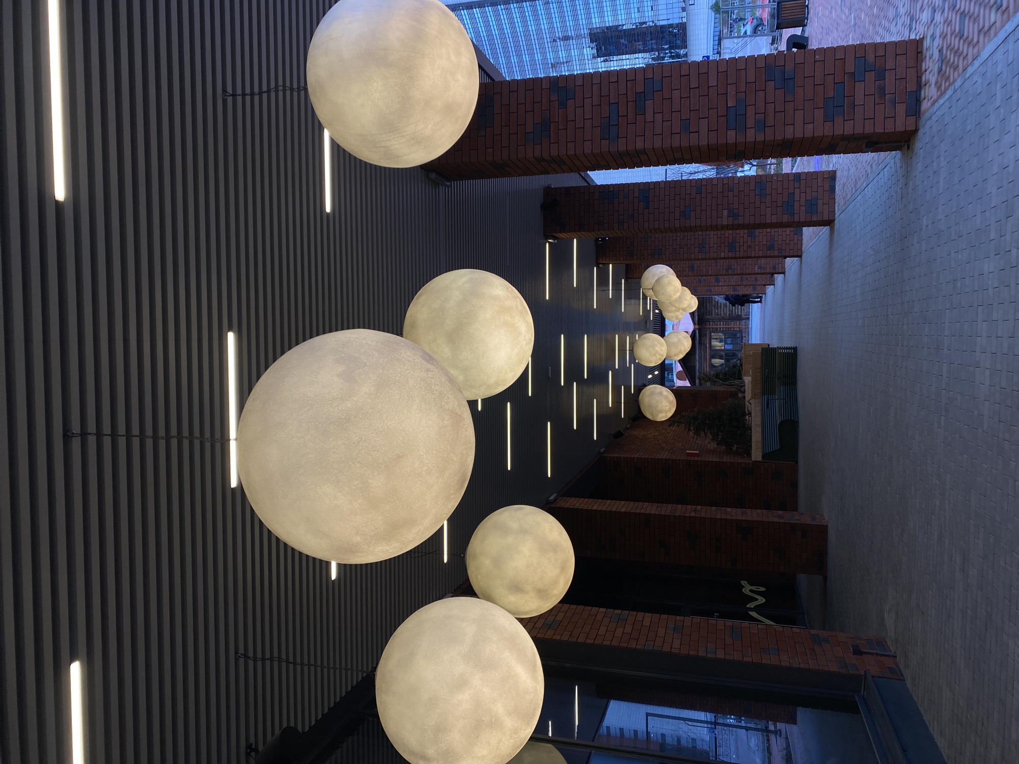 This image depicts a modern architectural space, likely a covered outdoor walkway or an atrium, at dusk or night in Yekaterinburg, Russia. The scene is dominated by numerous large, luminous spherical lamps, resembling moons, suspended at varying heights. These lamps glow with a soft, warm light and are arranged in a receding pattern, with larger ones in the foreground and smaller ones extending into the distance.
On the left side of the frame, a dark ceiling and adjacent wall are covered with vertical ribbed panels. Integrated into these panels are multiple bright, vertical strip lights that emit a cool white glow, contrasting with the warmer light of the spheres. To the right, a series of robust, rectangular structures clad in dark red and brown bricks extend horizontally overhead, suggesting supporting beams or architectural elements connecting surrounding buildings.
Below these brick structures lies a wide, light grey paved walkway, illuminated by the artificial lighting. A few small, indistinct figures of people can be seen strolling on this walkway in the distance, indicating the space is in use. The background reveals glimpses of modern urban buildings with reflective windows, further emphasizing the contemporary setting. The combination of the spherical lamps and strip lighting creates a serene and somewhat futuristic ambiance. No discernible text is visible within the image.