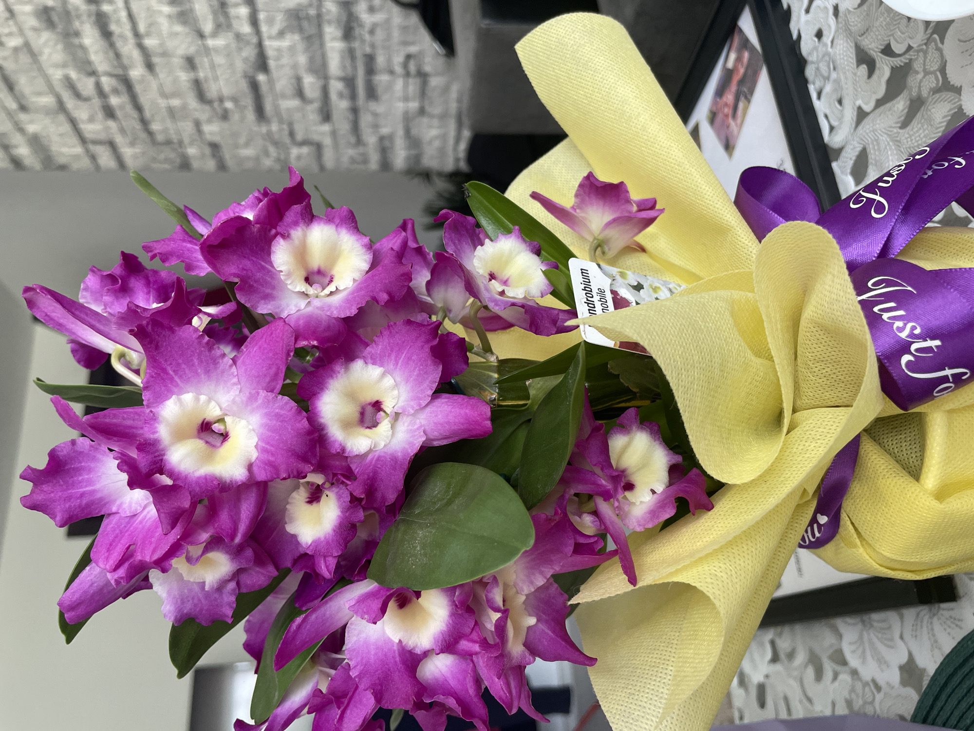 A bouquet of purple and white orchids is wrapped in yellow fabric and tied with a purple ribbon that says "Just for You". The background includes a white and grey brick wall, a black picture frame, and a white patterned tablecloth. The location is not discernible from the image.