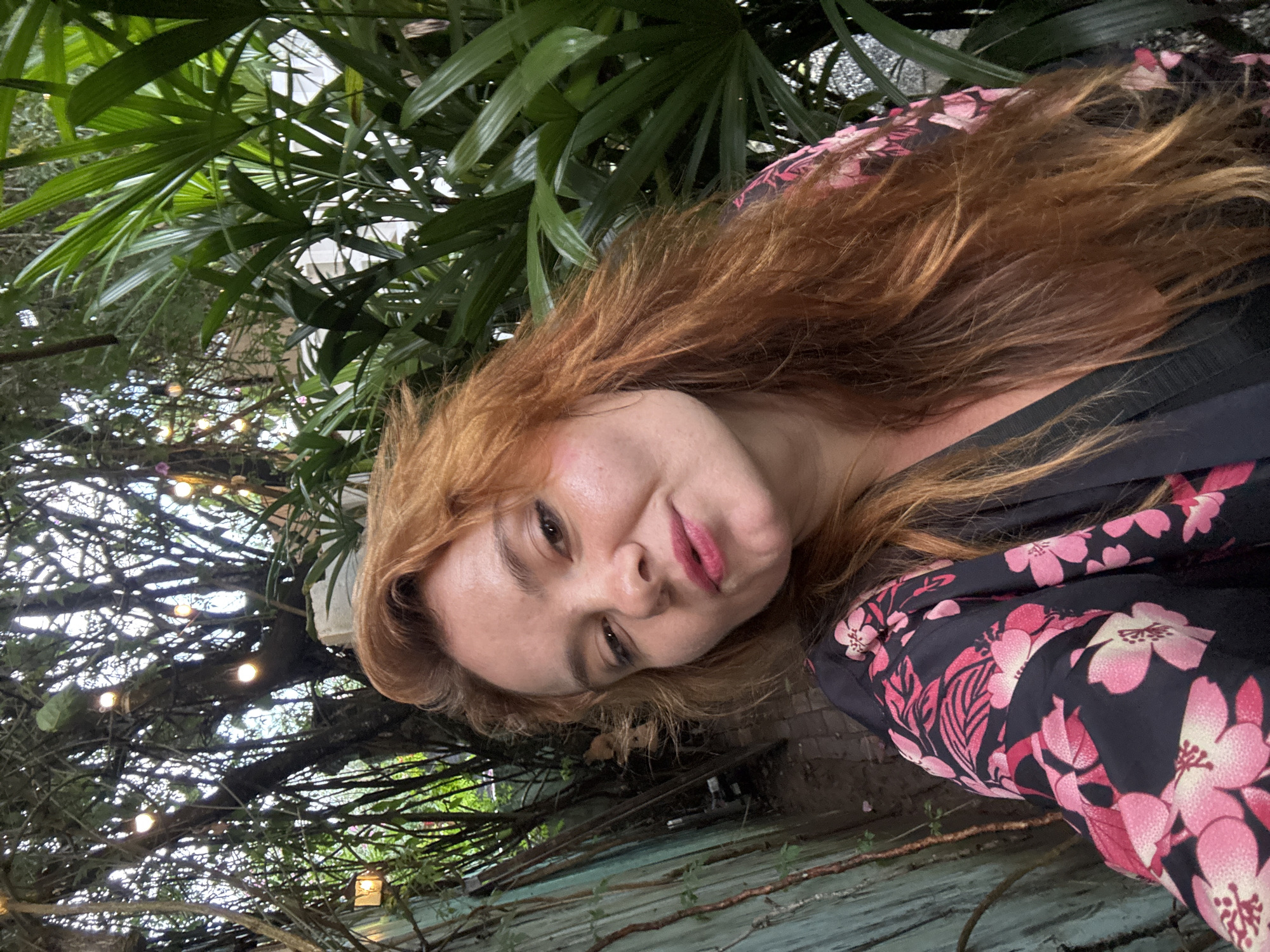 A young Caucasian woman with long, auburn hair and fair skin is pictured in a selfie. She is wearing a black robe with a vibrant pink floral pattern. Her head is tilted slightly, and she is looking towards the camera with a soft smile. The background is lush and green, with large tropical leaves on the right side of the frame. To the left, there are trees with numerous small, round, glowing lights strung between them, creating a festive atmosphere. A weathered, pale green wooden fence runs along the far left edge of the image. The ground appears to be paved with cobblestones. The lighting suggests it is either dusk or evening, as the artificial lights are prominent. This scene is consistent with the ambiance of many cafes and restaurants in Da Lat, Vietnam, known for their botanical gardens and fairy lights, often found in areas like the Xuan Huong Lake vicinity or the hillside resorts.