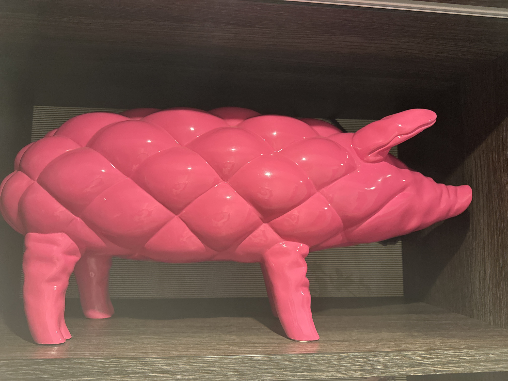 The image shows a bright pink, quilted-pattern pig figurine. It has a plump body with a pattern of raised, rounded squares resembling a quilted texture. The pig has short, stubby legs, a curled tail, and a protruding snout. It is placed on a shelf with a light-colored, wood-grain finish. Behind the pig, there is a striped pattern, suggesting a background of some kind. The lighting is even, highlighting the glossy finish of the figurine. The setting is simple and lacks any distinguishing features that would connect it to Genk, Belgium or any other specific location.