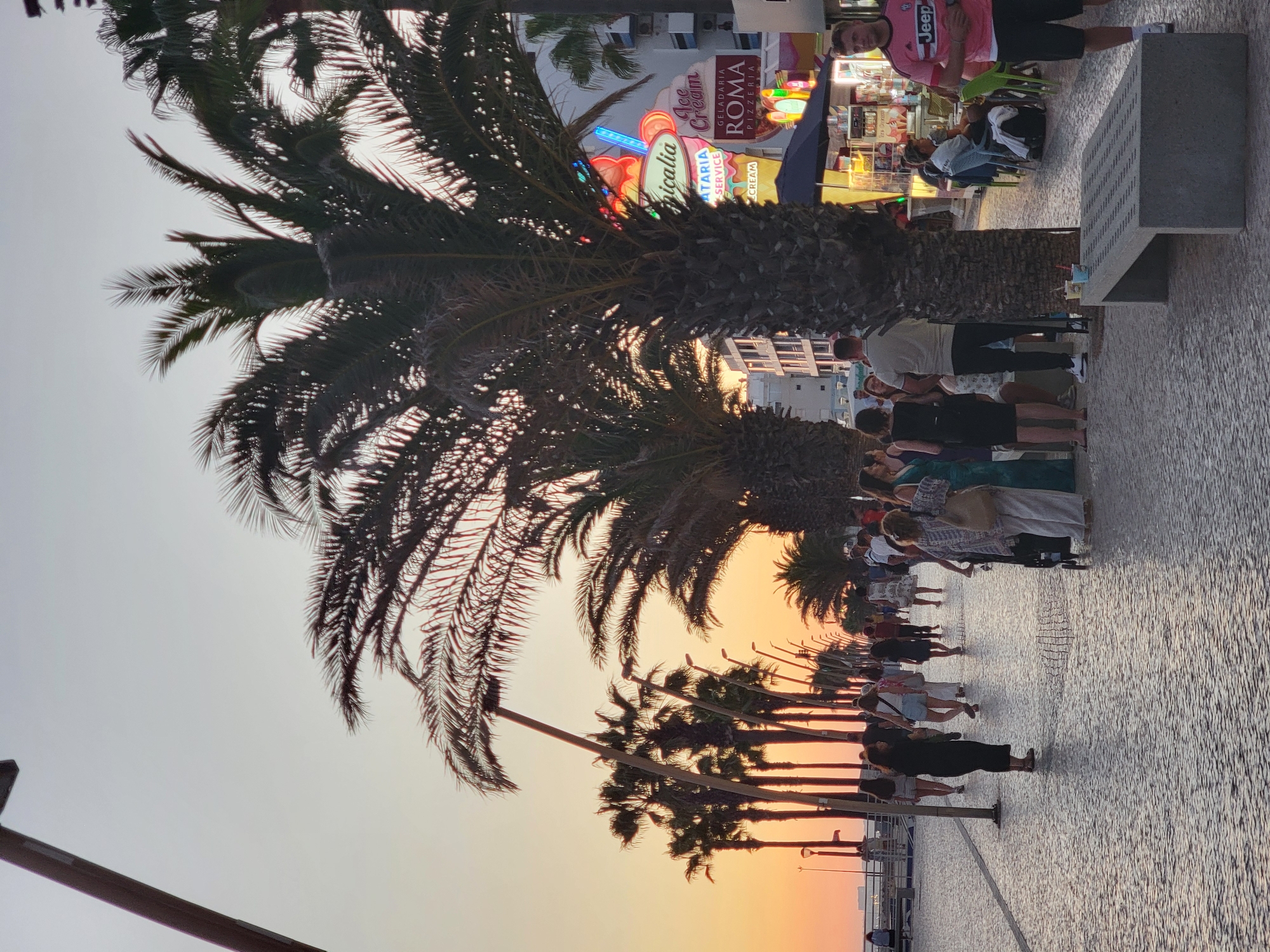 The image depicts a scene on the Quarteira promenade in Portugal during sunset. A paved walkway with a patterned stone surface is lined with palm trees on the right side. Several people are walking along the promenade, some in groups, and others individually. In the background, there are buildings with neon signs, including one that says "Tropicalia" and appears to be for an ice cream shop, and another that reads "Roma Geladaria Pizzeria." A man in a pink t-shirt with a "Jeep" logo is standing on the right side of the frame, looking towards the viewer. A concrete bench is positioned on the left side of the paved area in the foreground. The sky is a warm gradient of orange and yellow, indicative of sunset. The overall atmosphere is one of a lively evening gathering in a seaside town.