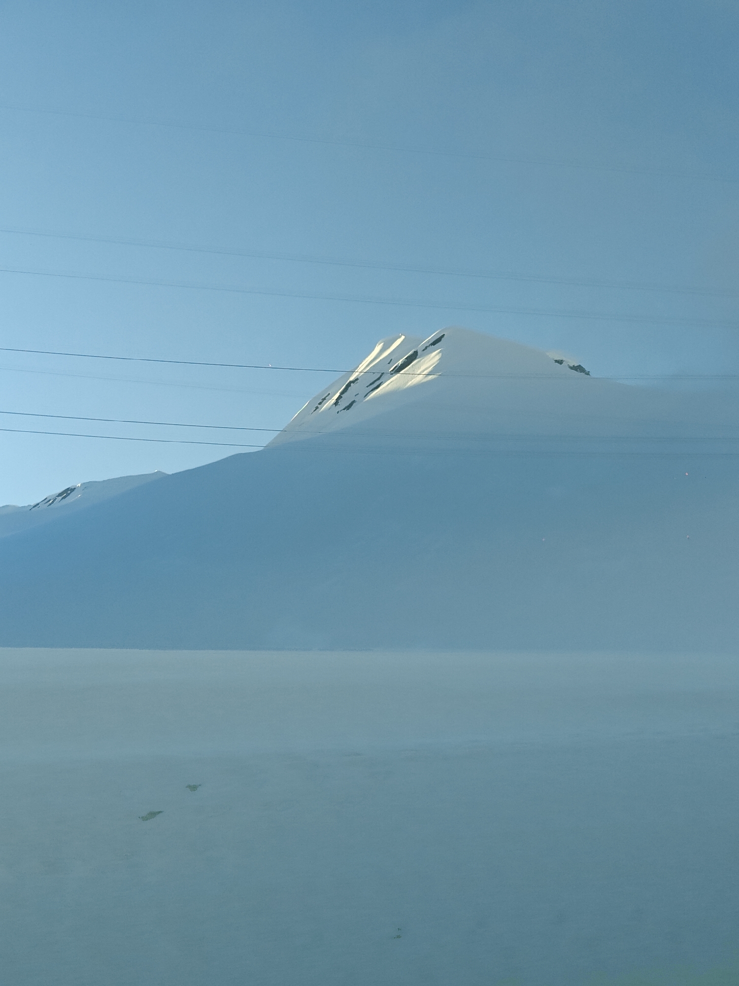 This image captures a vast, serene, snow-covered mountainous landscape in Ganisi, Georgia, under a clear, bright blue sky.
The prominent subject is a snow-capped mountain that dominates the upper and mid-ground. Its peak is brightly illuminated by direct sunlight, highlighting the pristine white snow and rocky outcrops. The lower slopes of the mountain appear partially obscured by shadows or a soft atmospheric haze, blending into the expansive, flat snowfield that stretches across the foreground and mid-ground. This snowfield is largely unbroken, creating a sense of immense scale and tranquility, with a few indistinct dark specks visible on its surface in the lower half of the frame, possibly small rocks or distant objects.
Several thin, dark utility lines traverse horizontally across the sky and mountain, introducing a man-made element into the otherwise natural scene. The sky is a uniform, vivid blue, suggesting a clear day. A faint, slender crescent shape, likely the moon, is visible high in the sky towards the upper right of the frame.
The time of day appears to be either morning or late afternoon, indicated by the angle of the sun brightly illuminating the mountain peak while leaving other areas in shadow. The weather is clear and sunny, implying cold temperatures typical of a snowy mountain environment. There is no visible text in the image. The overall impression is one of cold, quiet grandeur and expansive natural beauty.