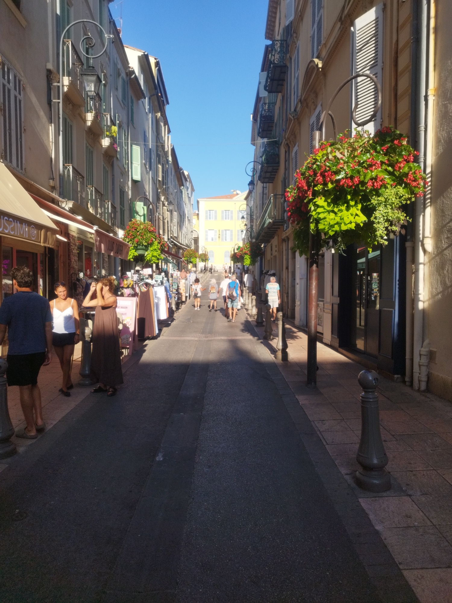 The image shows a sunlit street in Antibes, France, lined with buildings and shops. People are walking on the sidewalk and the street. The buildings have classic French architecture with shutters and balconies. Hanging flower baskets with red flowers add color to the scene. A large yellow building is visible in the distance. The street appears to be a pedestrian area with shops on both sides.