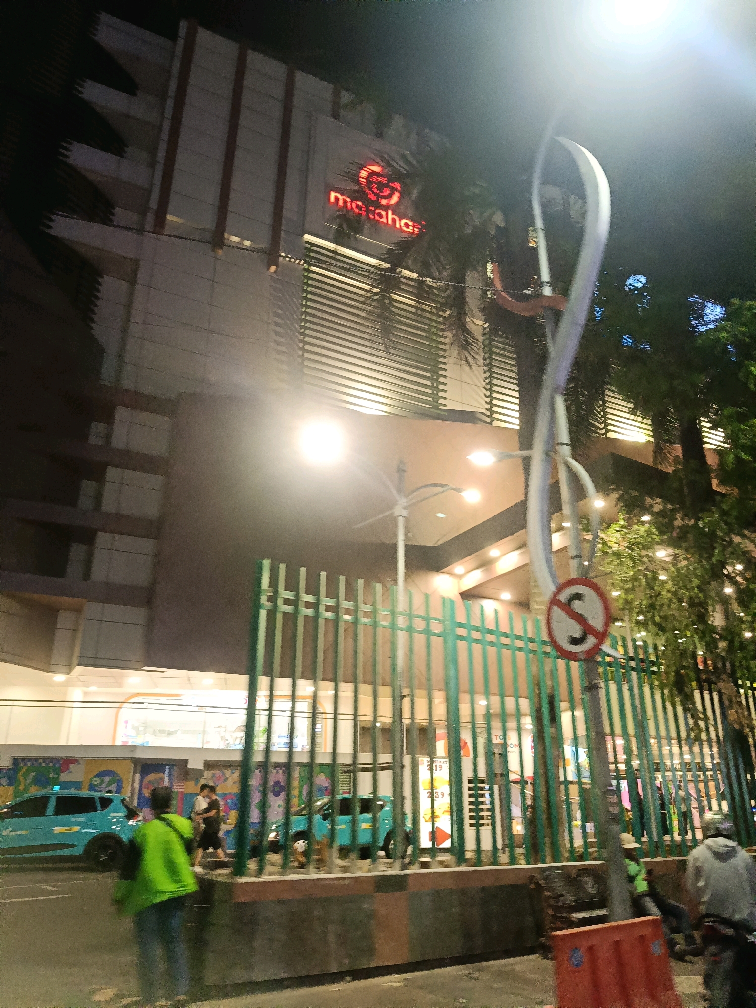 In the city of Surabaya, Indonesia, at night, a modern building with illuminated signs, possibly a department store or mall, stands prominently. The building's facade features horizontal louvers and is lit with warm yellow lights. A neon sign spelling "mataha" in red is visible on the upper portion of the building, likely part of a larger brand name. In front of the building, a green metal fence encloses the property.
On the street outside the fence, several elements are observable. Two light blue vehicles, possibly taxis or ride-sharing cars, are parked. A person wearing a bright green jacket and blue jeans walks away from the camera, facing towards the cars. Another two individuals are seen standing near the blue vehicles. A traffic sign, circular with a red border and a red diagonal line crossing over the letter "S" in black, is mounted on a pole near the fence. The street is paved, and there are visible light posts with multiple illuminated lamps. Trees with illuminated foliage are present, adding to the ambiance. The overall scene depicts a commercial area at night, bustling with activity and illuminated by artificial lights.