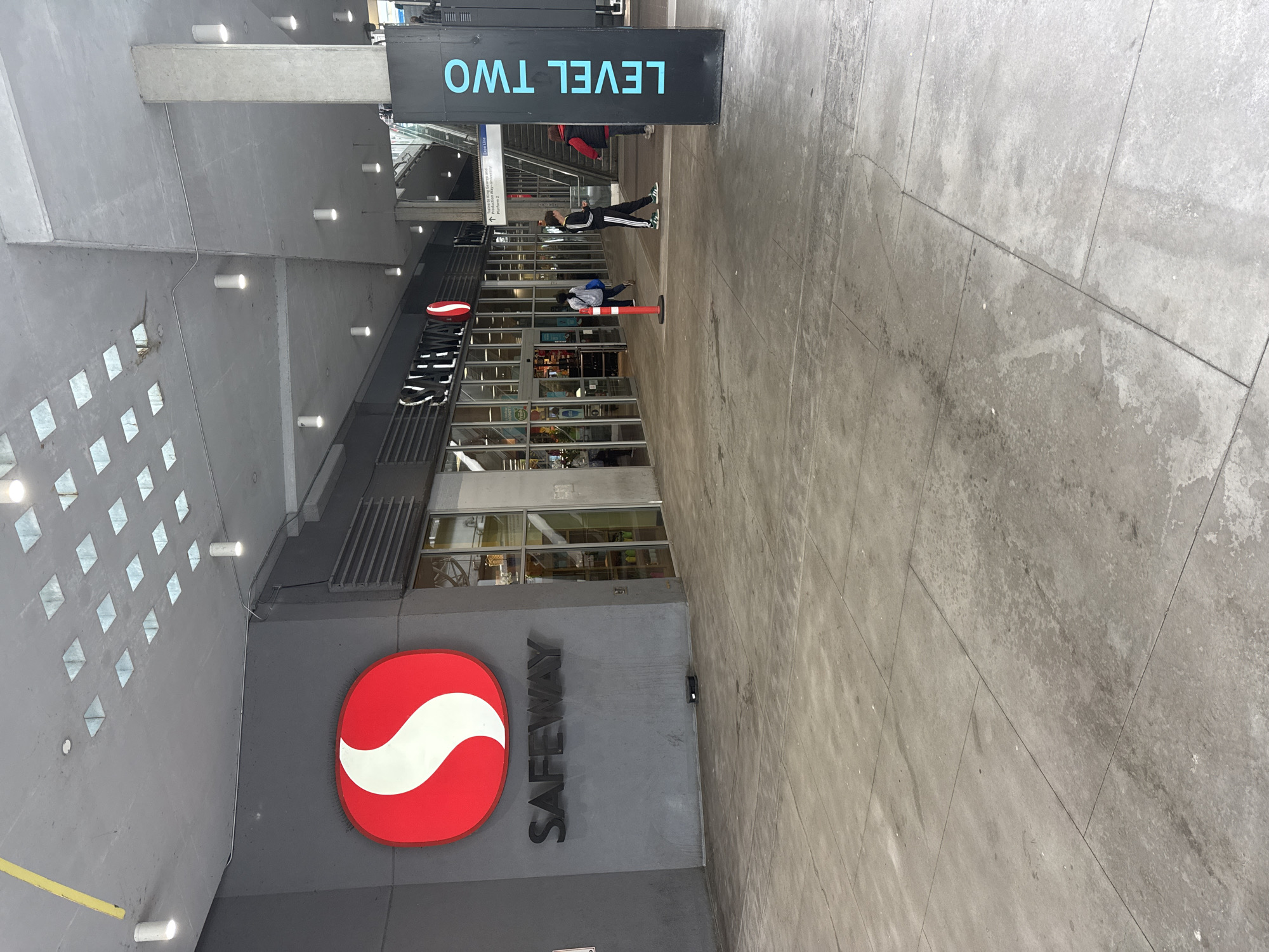 The image depicts the exterior entrance to a Safeway grocery store located in New Westminster, Canada. The store features a prominent red and white Safeway logo above the entrance. To the right of the store, a tall, black sign with bright blue lettering reads "LEVEL TWO". An escalator or staircase leading up to Level Two is visible behind the sign. Several individuals are present in the scene, including a person in a black tracksuit walking towards the camera and another person in a red vest near the staircase. The area is an outdoor, covered walkway with a concrete floor. The ceiling features a grid of square skylights.