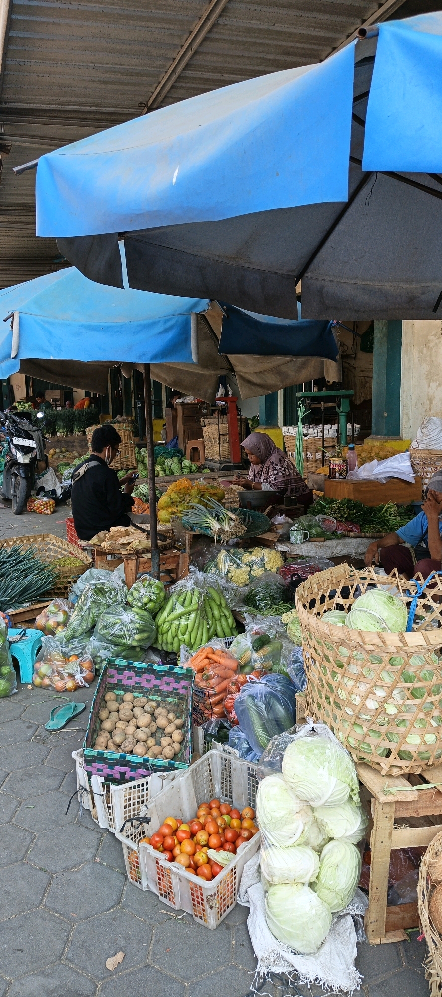 A street market in Klaten, Indonesia, displays a variety of fresh produce. Two individuals are visible; one, a person wearing a black shirt and face mask, is seated and looking at a phone. The other, a woman wearing a hijab, is seated and appears to be engaged in weighing produce. Stalls are covered by blue and grey tarps, offering shade. The ground is paved with hexagonal tiles.
The market is stocked with a wide assortment of vegetables and fruits, including cabbages, tomatoes, potatoes, carrots, cucumbers, watermelons, and green onions, many of which are packaged in plastic bags or displayed in baskets and crates. A motorcycle is parked to the left of the frame. A few small buildings with open doorways are visible in the background.