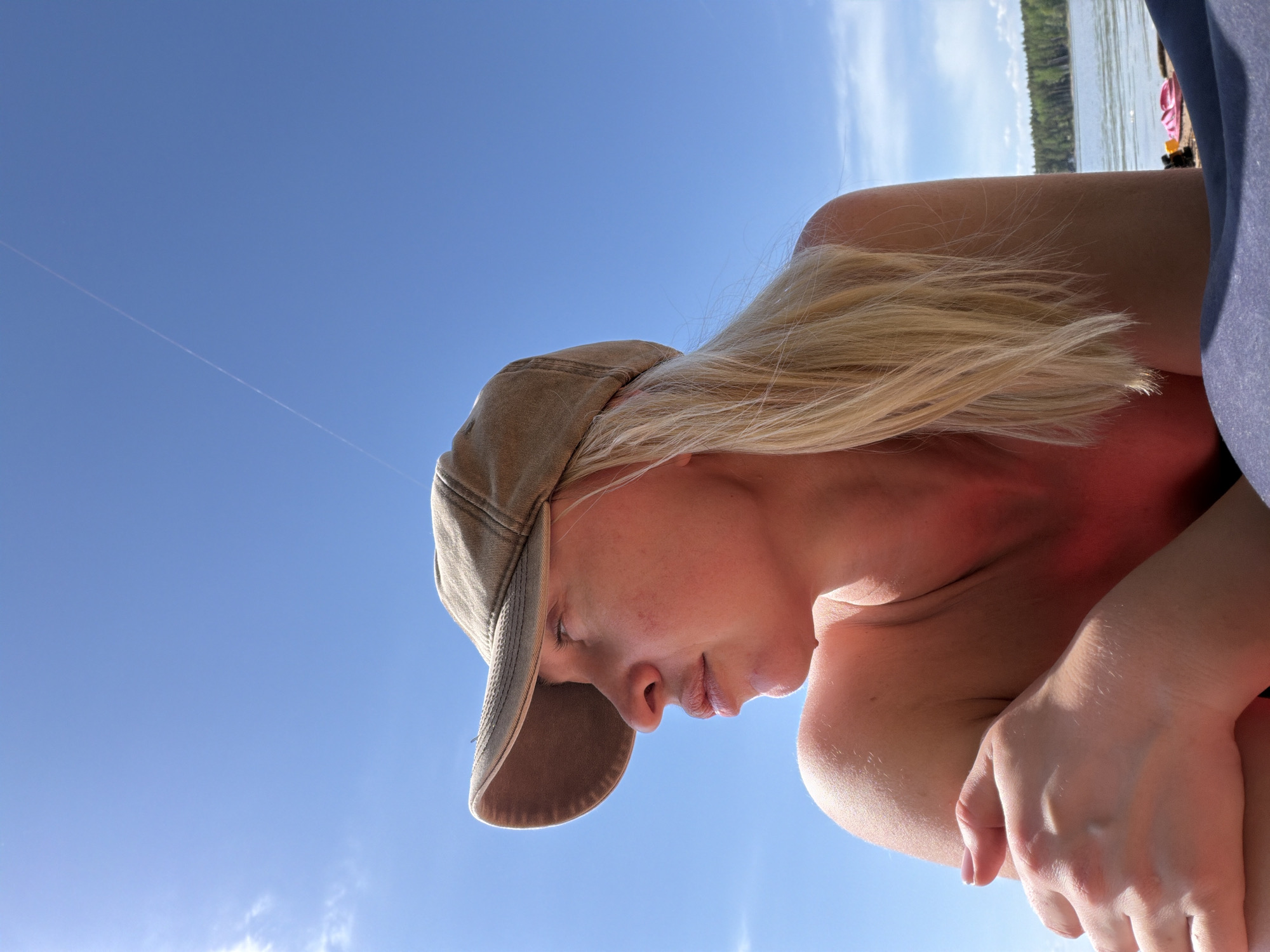 A low-angle shot depicts a fair-skinned person wearing a tan baseball cap. The background is a clear, bright blue sky. The person has blonde hair partially visible under the cap. The view includes the person's face in profile, neck, shoulder, and part of their arms. The environment includes what appears to be a lake and trees. Location: Zabegaevo, Russia.