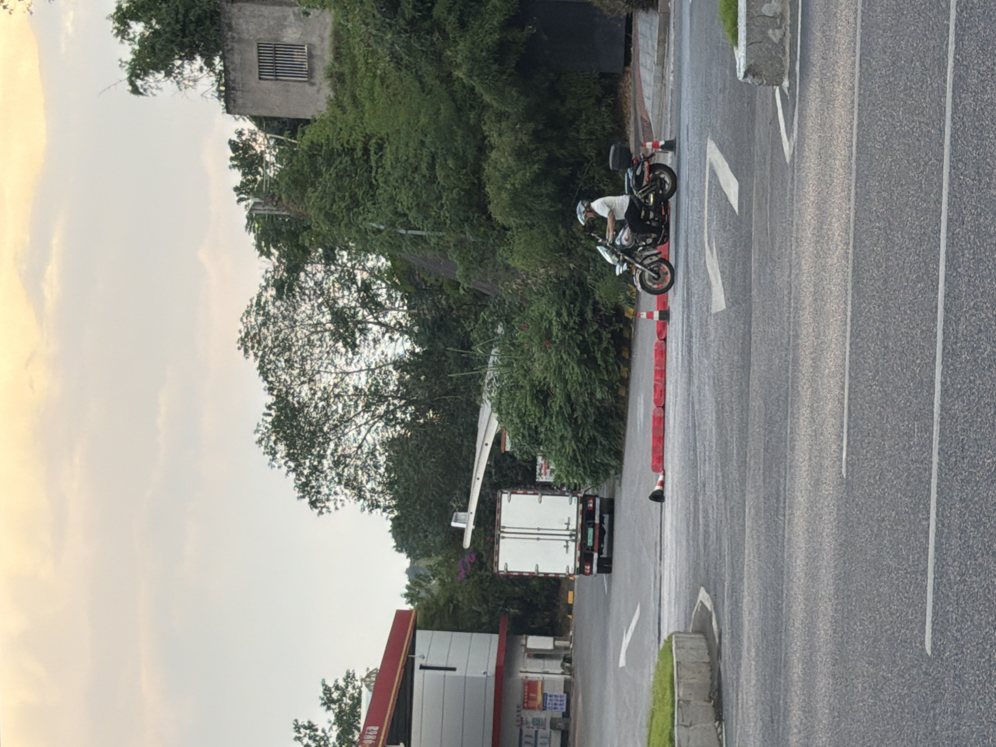 In Jiangjin, China, at a gas station, a white truck with a closed cargo box is parked on the right side of the road. On the left side of the road, a person is riding a motorcycle and is turning right. The sky is cloudy, and the sun is setting. The road is paved, and there are white lines on the road indicating lanes. There are also some traffic cones and barriers on the side of the road. The gas station has a red and white canopy. There are trees and bushes in the background, as well as a building on a hill.