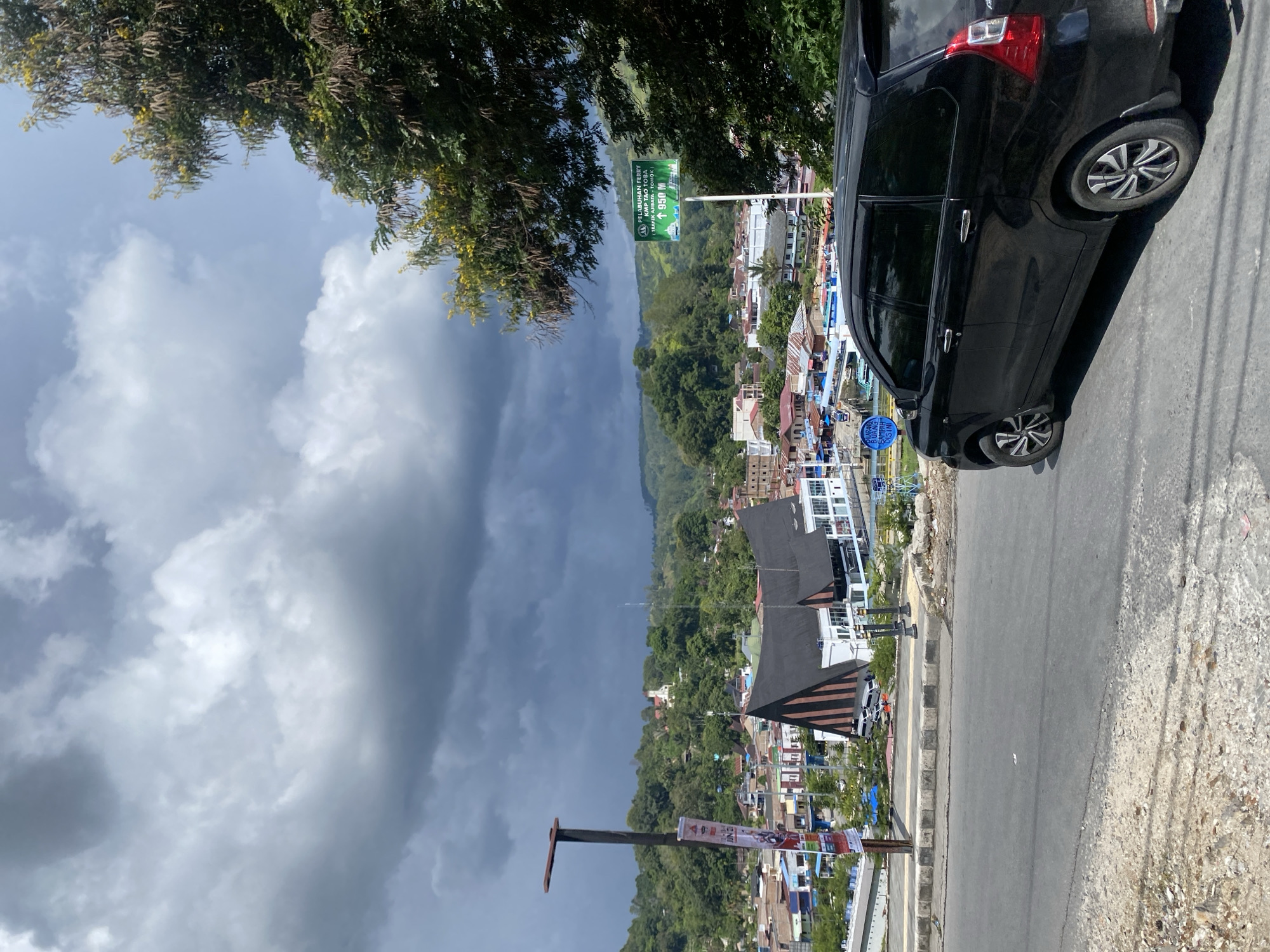 A black minivan is parked on a paved road overlooking a town nestled in a valley, with lush green hills and a dramatic, cloudy sky in the background. The town features a distinctive building with a black, angled roof and white walls, likely a public building or church. Other buildings in the town are of various colors and architectural styles. A green sign with white lettering is visible on the right side of the road, indicating "PELABUHAN FERRY" (Ferry Port) and a distance of 950 meters. A lamppost with a banner hangs on the left side of the road. The environment appears to be a scenic overlook in Parapat, Indonesia, possibly near Lake Toba, given the ferry port sign. The scene is devoid of people.