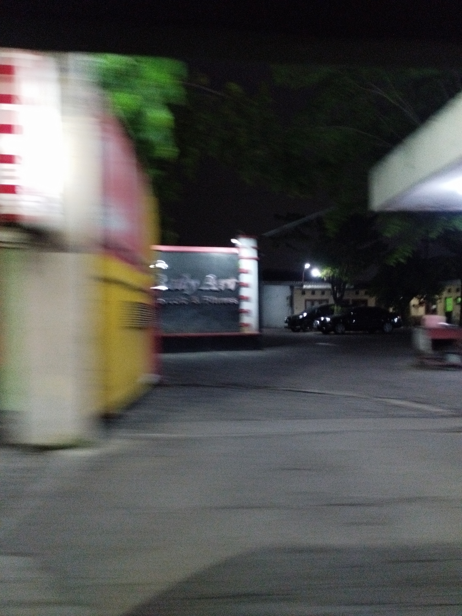 A blurred motion image captures a roadside scene at night, likely in the city of Surabaya, Indonesia. On the left, a brightly lit, partially visible structure with red and white stripes suggests a kiosk or shop. To the right of this, a dark sign with white text, possibly advertising "Daily Brew," is visible. Further to the right, several dark-colored cars are parked or moving on a paved road. The background features trees with green foliage and dimly lit buildings. The overall impression is that of a street or parking area with commercial activity, observed from a moving vehicle.