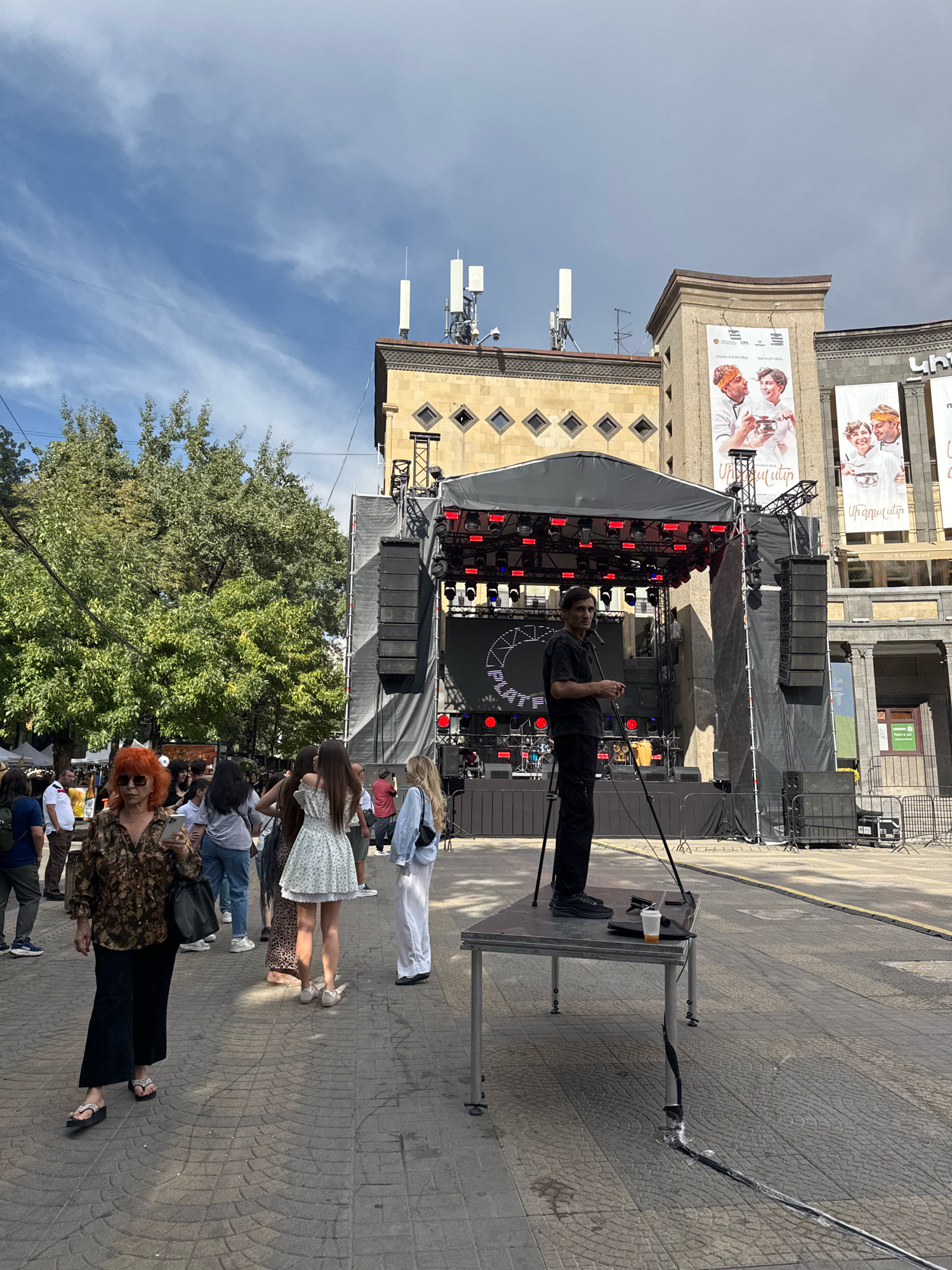 The image depicts an outdoor event in Yerevan, Armenia, likely a festival or concert, in front of a building with large banners featuring people. A stage with lighting and sound equipment is set up, draped in black fabric. A man in black attire stands on a metal table next to the stage, holding a tripod and looking towards the camera. Several people are walking on a paved walkway in the foreground, some are looking at their phones. Green trees are visible in the background, and the sky is partially cloudy. The architecture of the building suggests a public space or commercial area in the city.