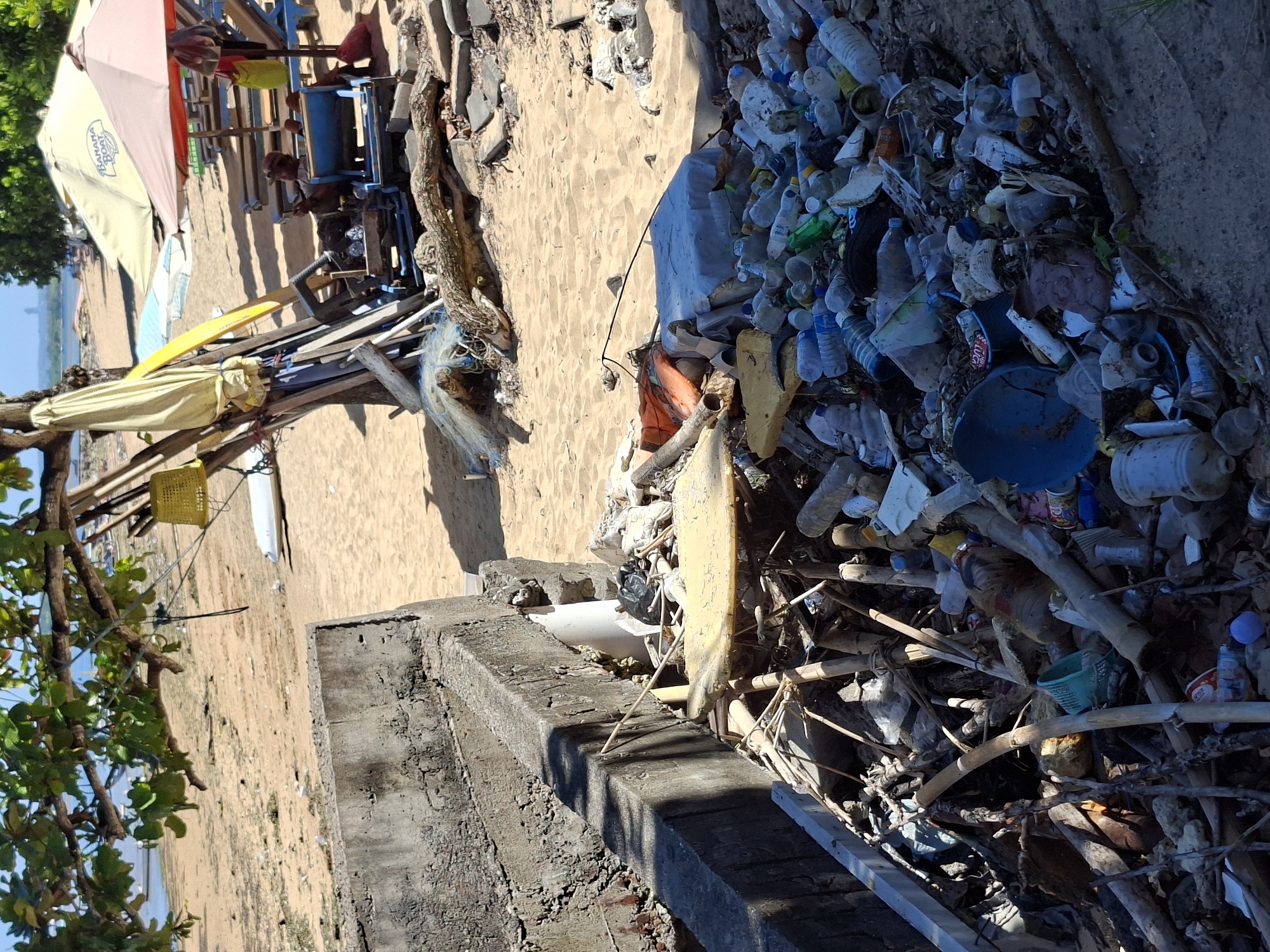 The image depicts a heavily polluted beach in Denpasar, Indonesia. A large accumulation of plastic waste, including bottles, containers, and miscellaneous debris, is concentrated near a concrete structure, possibly a retaining wall or steps leading to the beach. Beyond the debris, the sandy beach stretches out towards a small collection of makeshift structures, sun umbrellas, and beach furniture. Some of these items appear to be in a state of disrepair or abandonment. In the background, one can see the ocean meeting the horizon under a clear sky. A tree with green foliage occupies the lower left foreground, partially obscuring the scene.