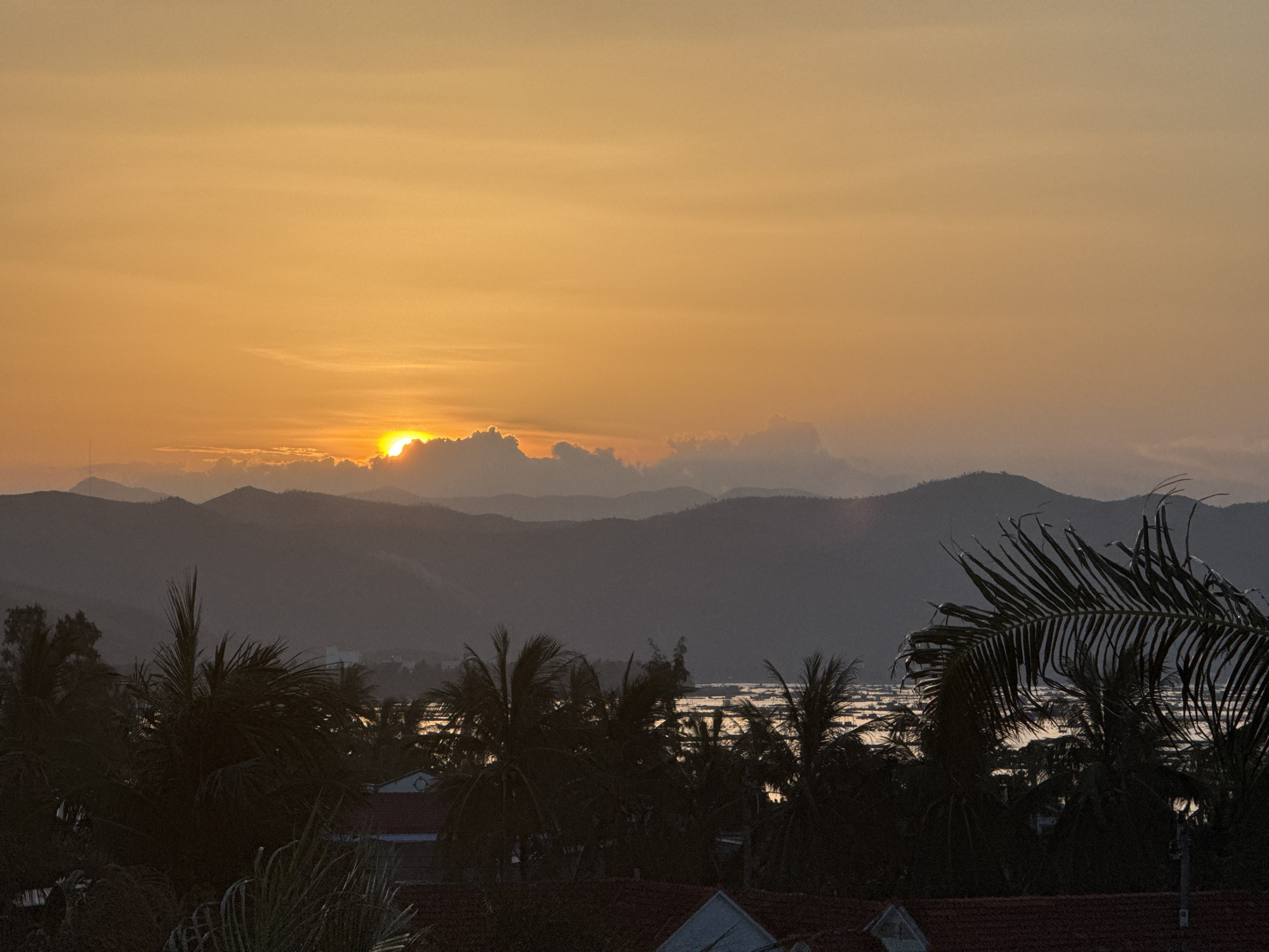 This image captures a sunset over the city of Song Cau, Vietnam. The sky is a vibrant orange, with the sun partially obscured by clouds on the horizon. In the foreground, silhouetted palm trees and rooftops are visible, hinting at a tropical landscape. Beyond the immediate trees, a body of water reflects the warm glow of the sunset, with what appear to be small structures or boats dotting its surface. The background is dominated by layers of hazy mountains, fading into the distance. The overall impression is one of tranquility and natural beauty as day transitions into night.