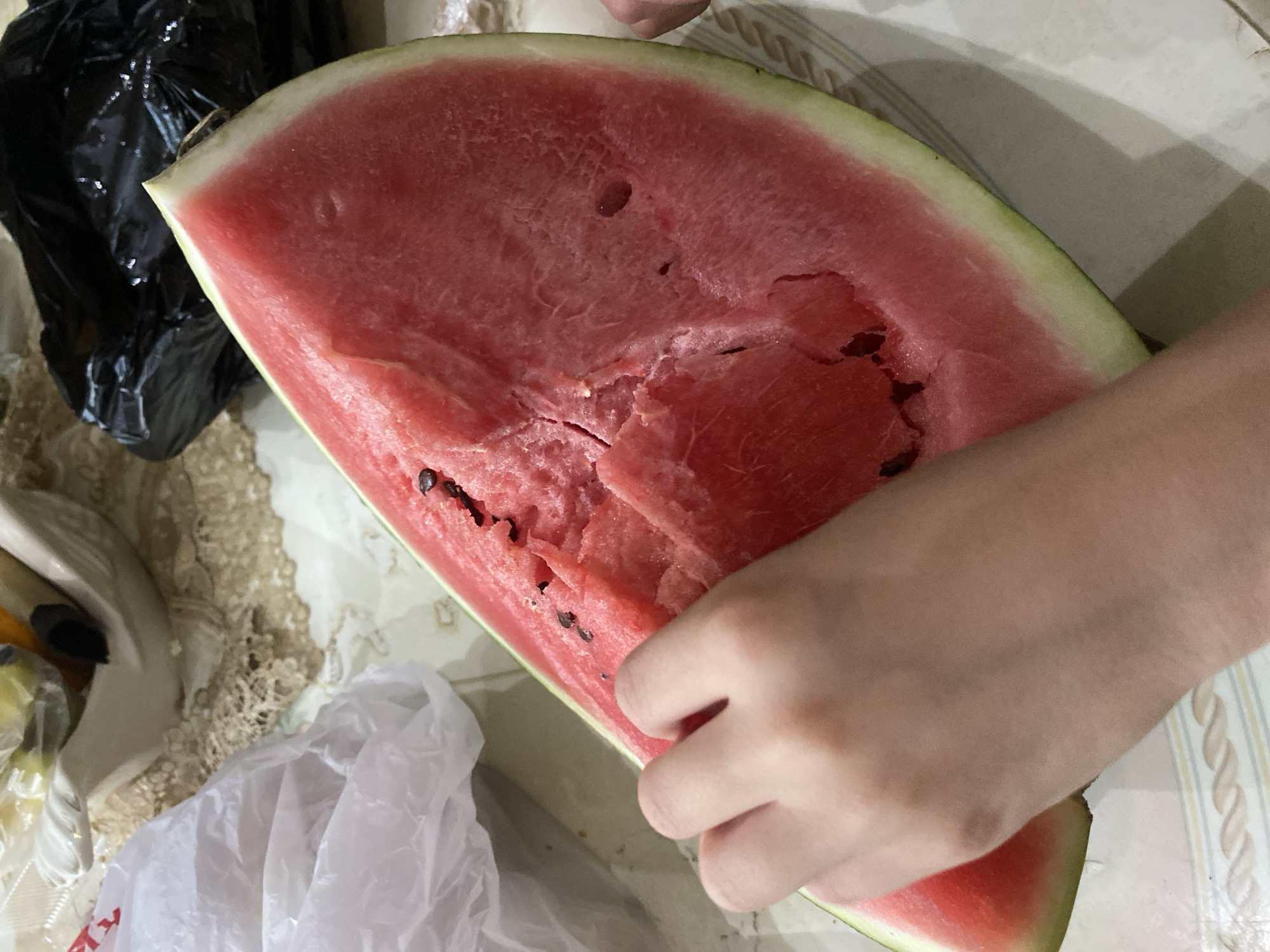 The image shows a large slice of watermelon with some of its flesh eaten away. A person's hand is visible, touching the watermelon. The watermelon is placed on a patterned surface, likely a table covered with a tablecloth. In the background, there are plastic bags and a decorative object. There is no specific information in the image that allows for the determination of its location in Volzhsk, Russia.