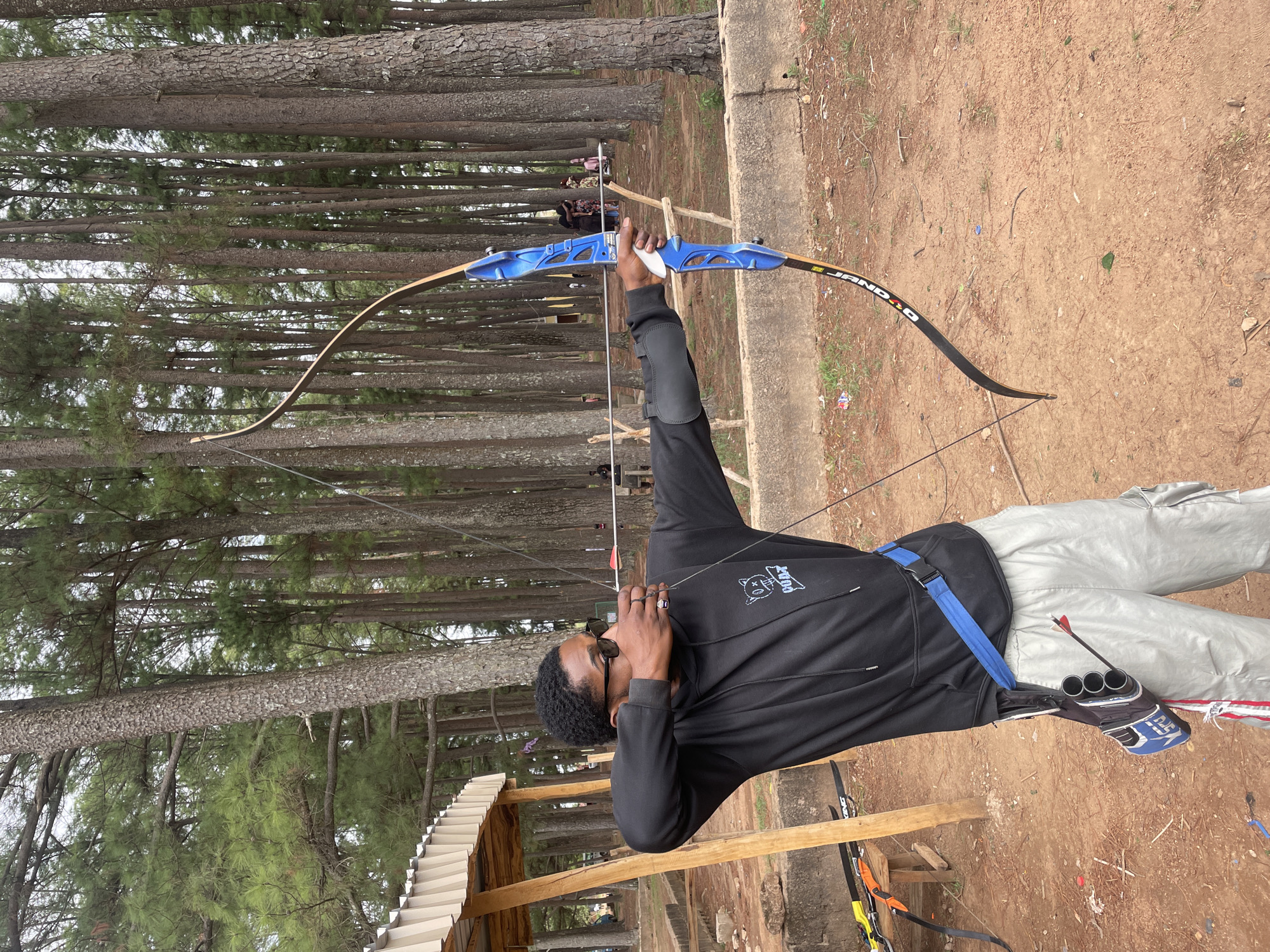A man is drawing back a recurve bow at an archery range. He is wearing a black hooded sweatshirt, light grey cargo pants, sunglasses, and a forearm guard. The bow is blue and black, and he is pulling the string back to his face. Arrows are visible in a quiver on his hip. The archery range is located in a wooded area, with tall pine trees in the background. The ground is dirt and pine needles. A partially constructed wooden structure with a corrugated roof is visible in the lower left corner. The location is Jos, Nigeria.