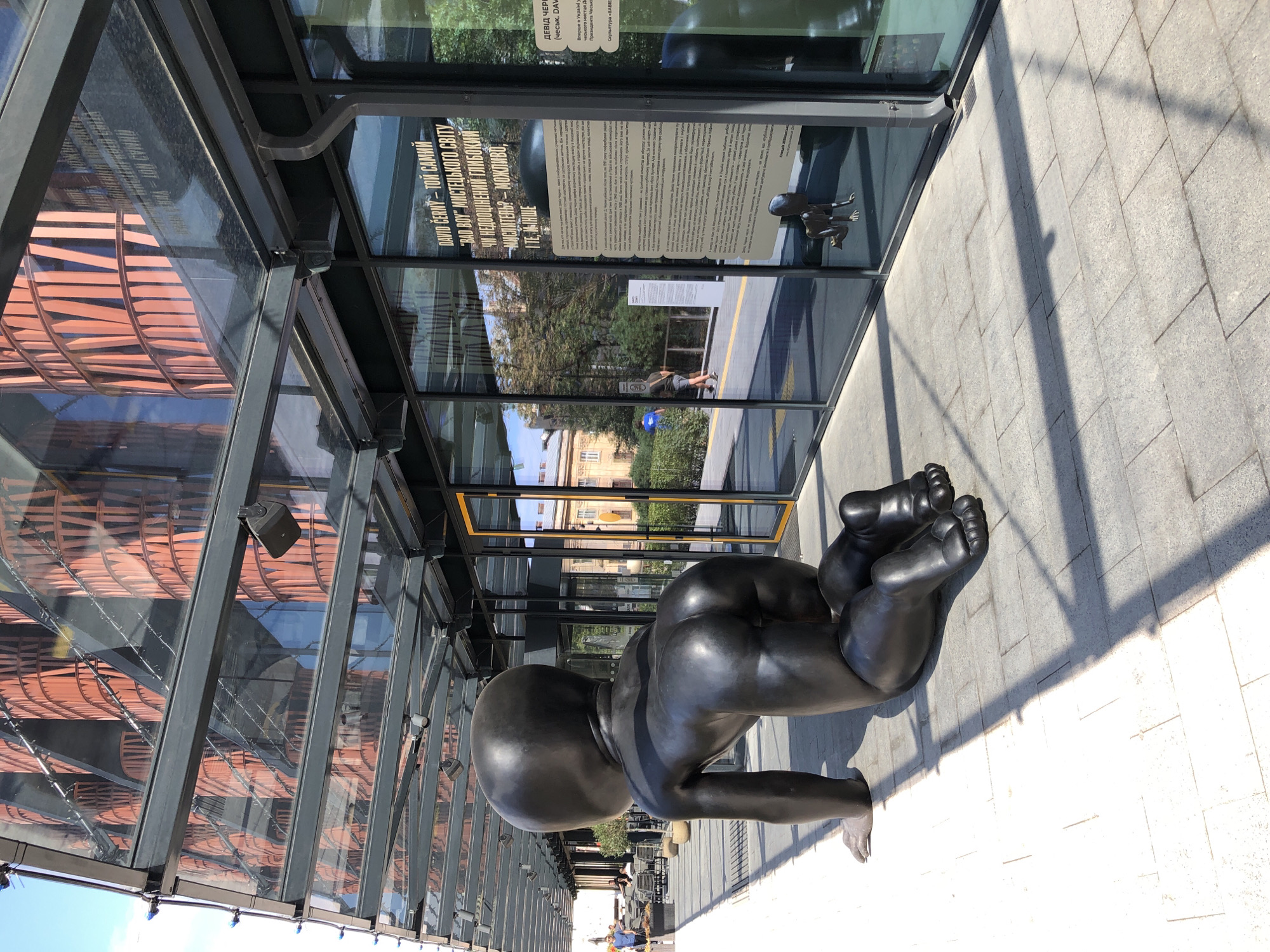 The photograph shows a large, dark sculpture of a crawling baby in an outdoor setting, likely a public space or an exhibition area. The sculpture is positioned on a paved walkway, with its back facing the camera. The baby's head is large and disproportionate to its body, a stylistic feature of the artwork.
In the background, a modern building with a facade of orange, vertical elements and a glass-covered walkway is visible. The glass reflects the building and the sky, creating a shimmering effect. To the right, large glass windows display text in Ukrainian, suggesting an exhibition or informational signage related to the sculpture or the building. A smaller, similar sculpture of a crawling baby is visible reflected in one of the windows.
The environment appears to be a sunny day, casting shadows from the glass structure and the sculpture onto the ground. In the distance, to the left, some people are seen sitting at tables outdoors, and others are walking along a pathway. The overall scene suggests an urban environment, possibly a cultural or artistic space in Lviv, Ukraine, given the presence of the Ukrainian text on the signage and the architectural style.