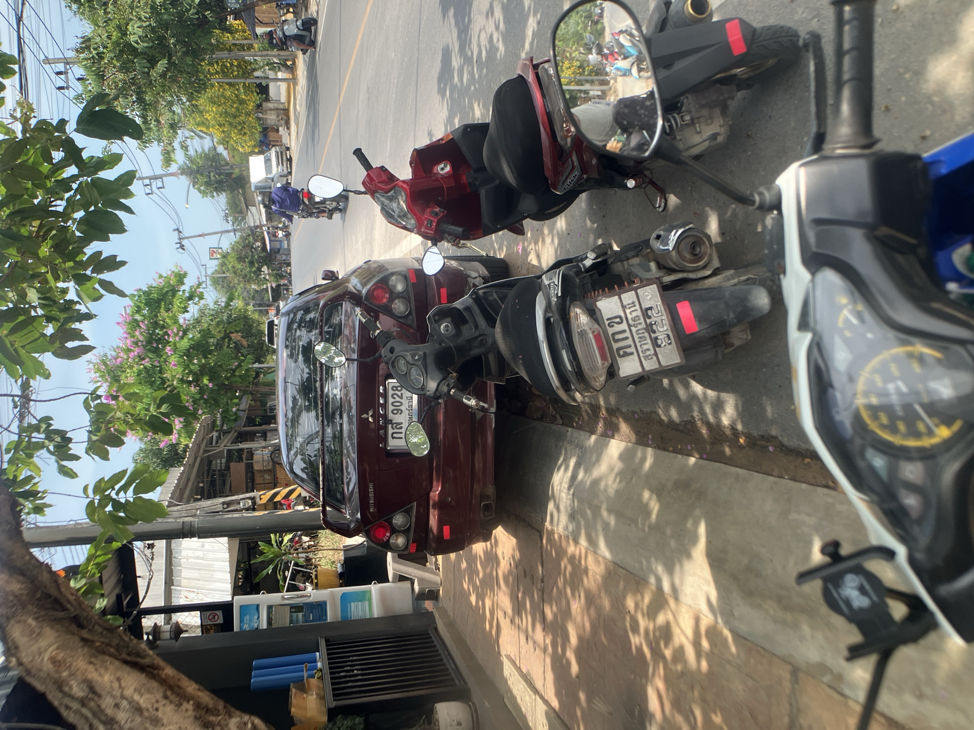 The image depicts a street scene in Baan Plai Laem, Thailand. A maroon Mitsubishi Lancer is parked on the side of the road, next to a motorcycle with license plate "กล 9028 สุราษฎร์ธานี" and another motorcycle with license plate "คกข 25 สุราษฎร์ธานี." A third motorcycle is partially visible on the right edge of the frame. Trees and buildings are visible in the background, and shadows are cast on the street.