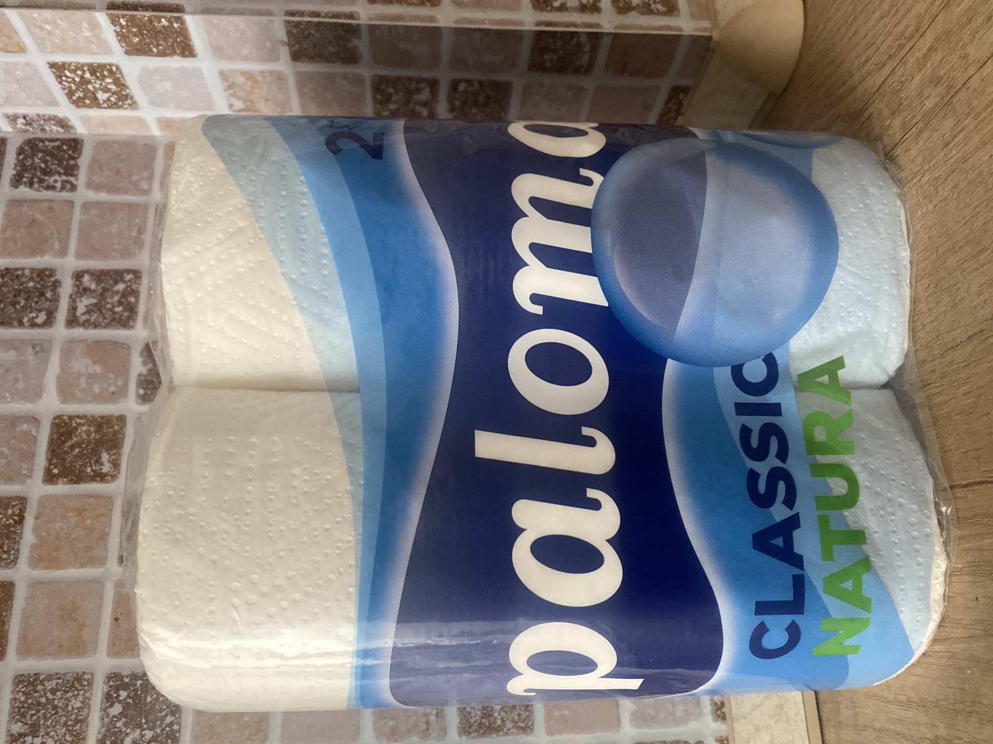 A plastic-wrapped package of Paloma Classic Natura toilet paper is positioned on a wooden surface in front of a tiled wall. The wall has a mosaic pattern of square tiles in various shades of brown, beige, and white. The toilet paper package is predominantly blue with white and light blue wave-like graphics. The brand name "paloma" is prominently displayed in large white cursive lettering. Below that, "CLASSIC" is written in dark blue capital letters, and "NATURA" is in green capital letters. The package contains two rolls of toilet paper, visible through the transparent plastic. A blue, semi-transparent sphere graphic is superimposed on the lower right portion of the package. The lighting suggests an indoor environment. There are no people or animals visible in the image, and no specific landmarks or identifiable features that would suggest a location in Chișinău, Moldova.