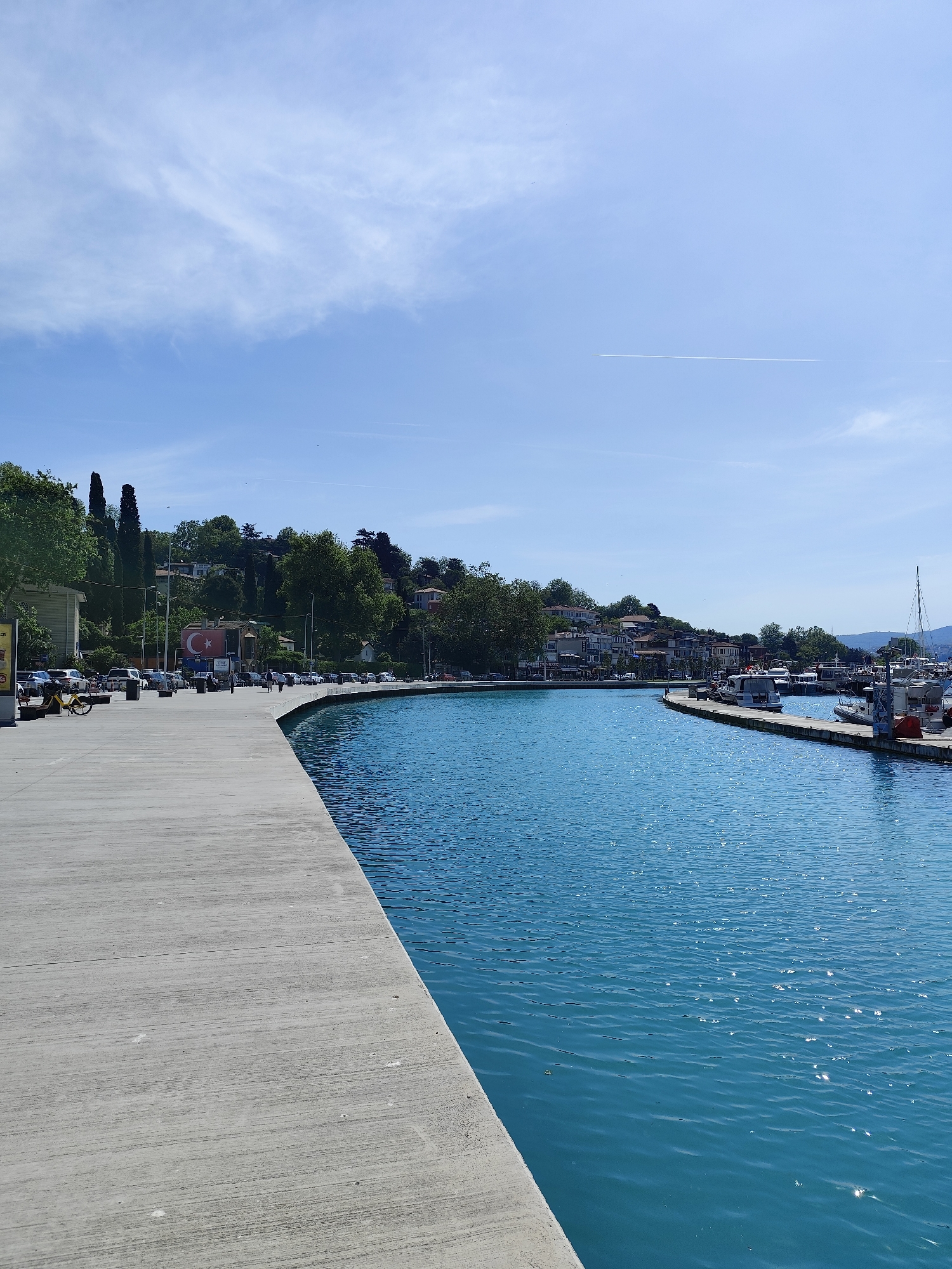 The image shows a sunny day in Sarıyer, Turkey. A concrete walkway curves along the edge of a body of water with boats docked nearby. Across the water, buildings are nestled among trees on a hillside. A Turkish flag is visible on one of the buildings. Cars are parked along the road adjacent to the walkway, and several people are walking. The sky is blue with scattered clouds.