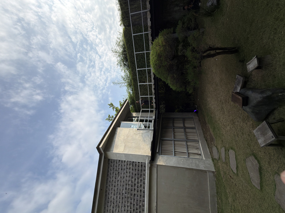 This image depicts an outdoor scene in Tumbak Bayuh, Indonesia, featuring a modern architectural structure and a landscaped garden under a partly cloudy sky.
The primary subject is a two-story building constructed with grey concrete and a section of textured grey brickwork on its upper facade. A dark roof covers the structure. On the upper level, a glass door opens onto an elevated, flat walkway or bridge with grey metal railings. One individual, dressed in dark attire, is standing on this walkway, facing left. The lower level of the building features a wall with large, grid-framed windows, behind which light-colored curtains are visible.
The ground area in front of the building is a green lawn. A winding path of irregular grey stone stepping stones traverses the grass towards the building. In the foreground, several rustic outdoor furniture pieces are present: two square wooden stools and a larger dark, irregular base (possibly stone or concrete) topped with a rectangular wooden box. A small, gnarled tree with green foliage stands on the right side of the lawn. Beyond the elevated walkway, a dense hedge of green foliage forms a boundary, with taller trees and bamboo visible above it. On the far right, partially obscured by vegetation, a second individual is visible, seated or crouching on the grass, wearing dark clothing.