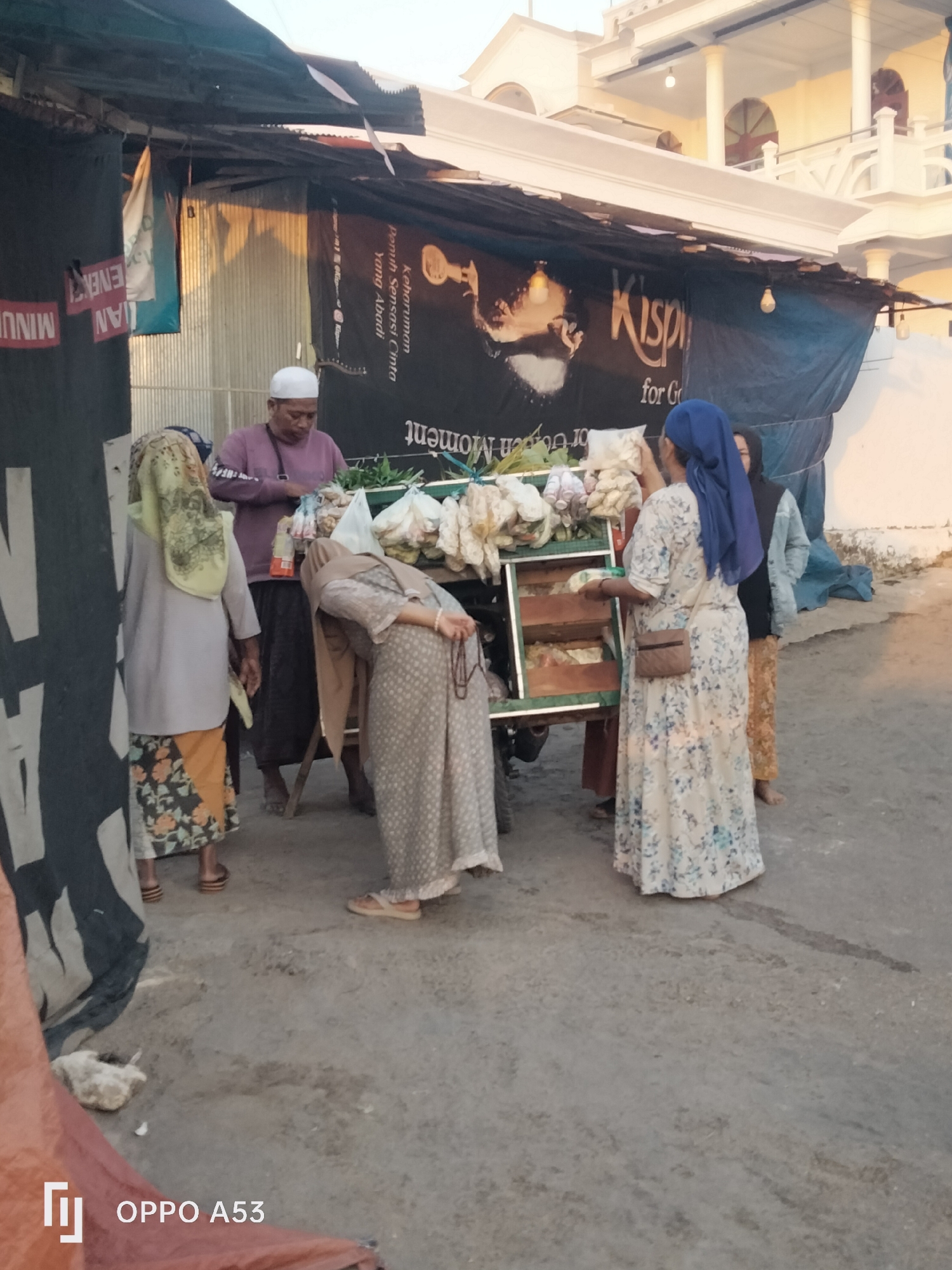 In Badung, Indonesia, a street vendor displays produce from a cart under an awning. A man in a white kufi and a purple shirt stands behind the cart, with several women, all wearing hijabs and modest clothing, browsing the goods. One woman in a blue hijab selects items, while another in a patterned dress bends over the cart, and a third in a light gray garment and floral skirt stands to the side. The scene is illuminated by the warm glow of the late afternoon sun, casting shadows on the ground and highlighting the textures of the corrugated metal wall behind the stall. A large banner with Indonesian text and an image of a face hangs above the vendor's setup. The ground is paved with rough, grey concrete.