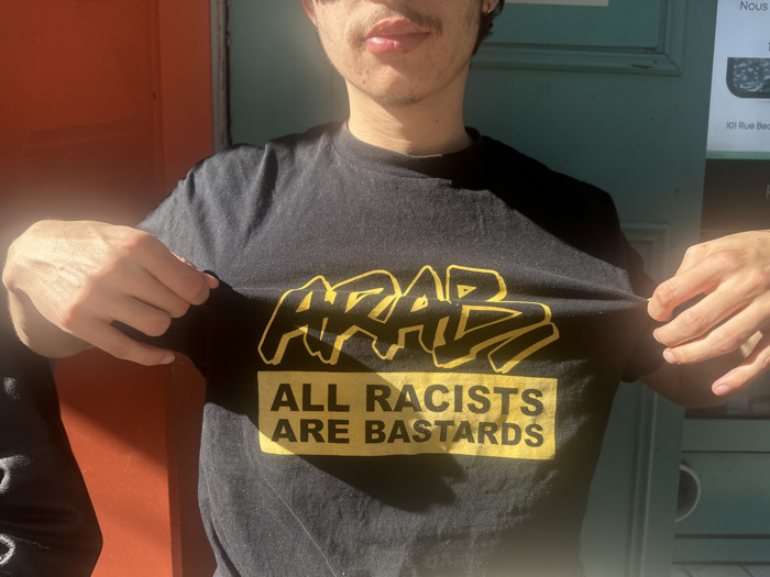 A male individual is captured from the chest upwards, standing outdoors in direct sunlight. He is wearing a black short-sleeved t-shirt with a dual-line yellow inscription. The upper line, in a stylized graffiti-like font, reads "ARAB." Below this, within a yellow rectangular block, the text "ALL RACISTS ARE BASTARDS" is displayed in a sans-serif font. The individual uses both hands to pull the t-shirt outward, ensuring the text is clearly visible. His facial features include a visible mustache and chin hair; a small gold earring is discernible in his left earlobe. The background comprises two vertical structures: a reddish-orange wall or door on the left and a faded teal-green door or shutter on the right. A white sign affixed to the teal-green surface is partially visible, displaying the text "Nous" and "101 Rue Bec," which indicates a specific location in the city of Paris, France.