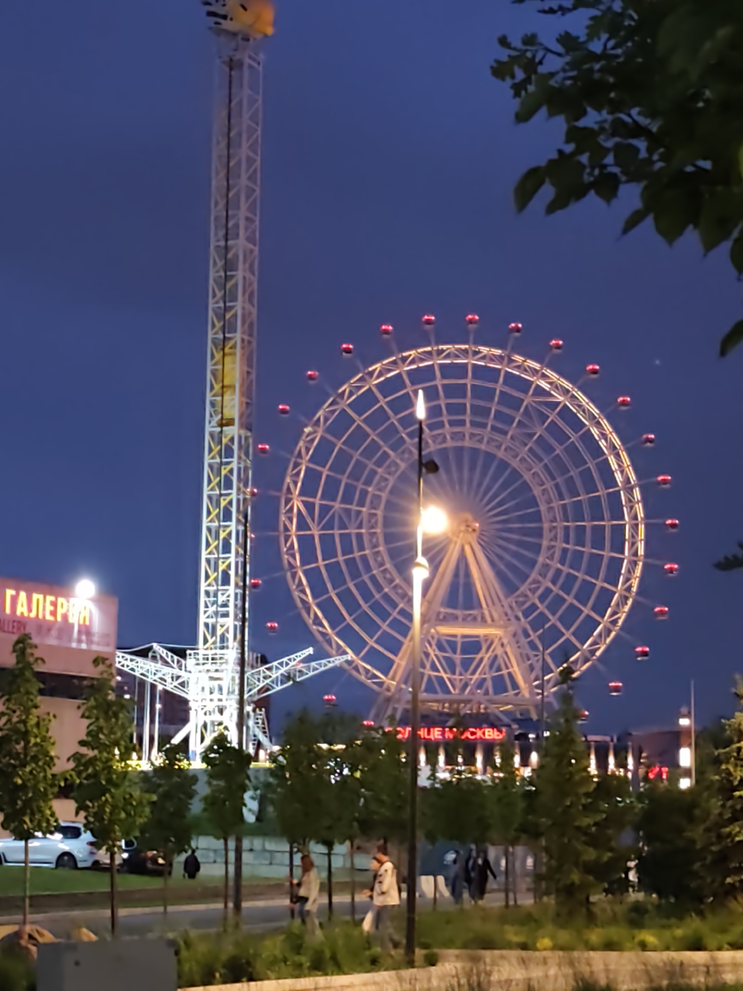 The image captures an amusement park scene in Moscow, Russia, at dusk or early evening. A tall drop tower stands to the left, illuminated with white lights. Adjacent to it is a large Ferris wheel with red and white lights. The base of the Ferris wheel is partially obscured by trees and a streetlight.
In the foreground, people are walking along a sidewalk lined with trees. There is a building with the word "ГАЛЕРЕЯ" (Gallery) visible on it. Cars are parked along the street. The overall atmosphere is that of a public leisure area at night. The Ferris wheel and drop tower are the main points of interest, likely attractions within the park.