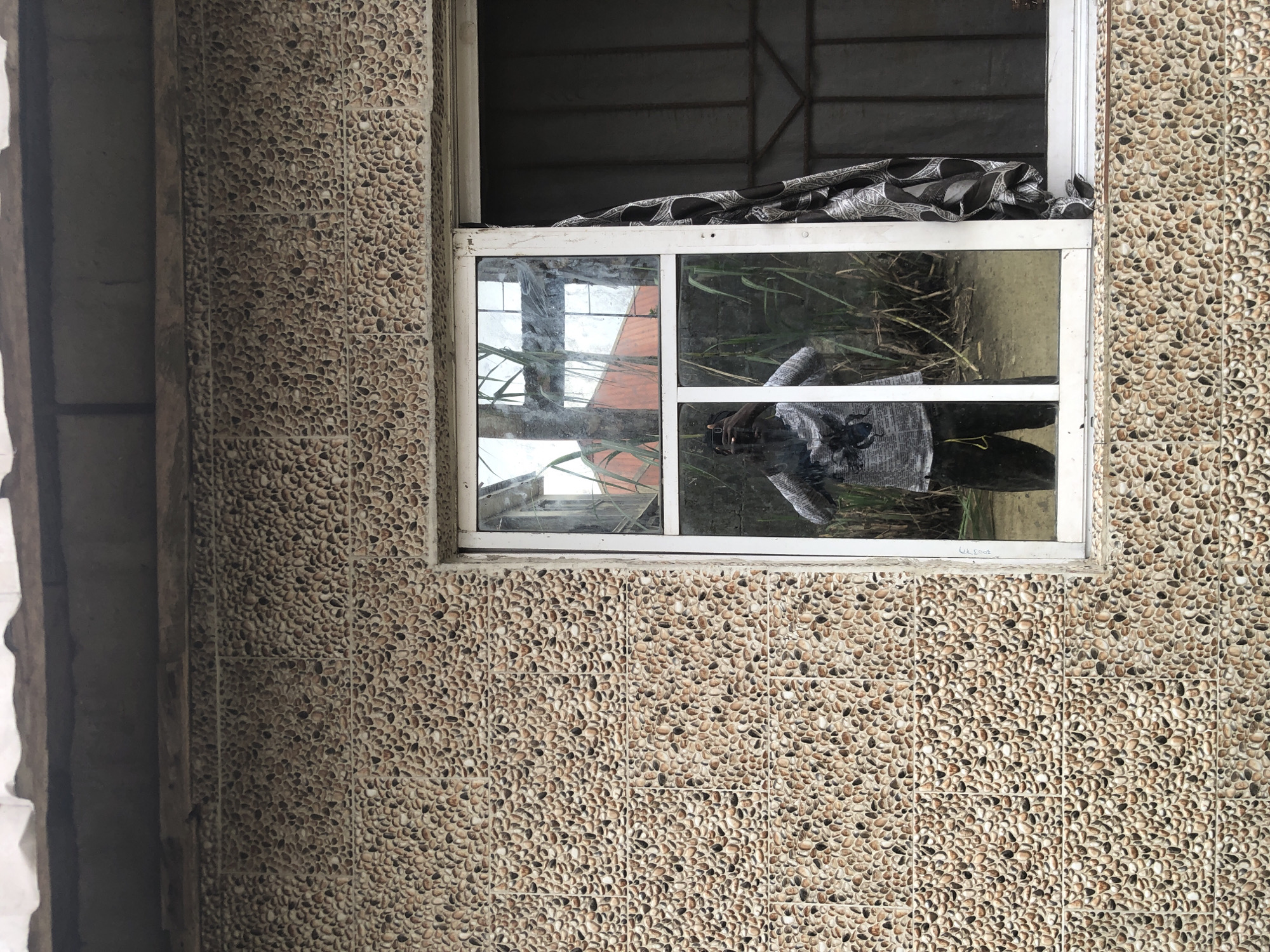 A person is reflected in a window, holding a phone to take a photograph of themselves. They are wearing a patterned shirt and dark pants. The window is set in a wall covered in small, rounded stones of various brown and tan colors. A light-colored curtain with a black and white pattern hangs on the right side of the window. The reflection shows greenery and a reddish building in the background. The image appears to be taken outdoors, possibly in a residential area in Ado, Nigeria.