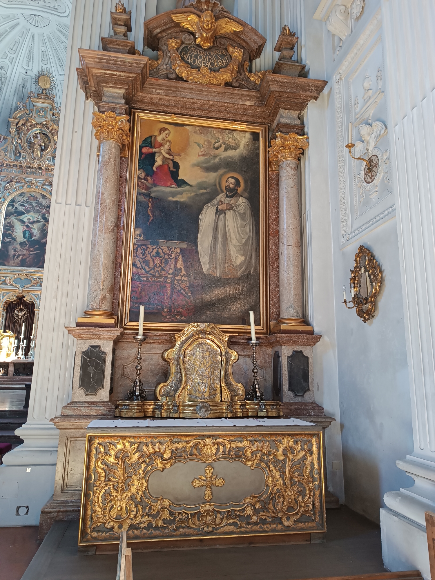 The image shows an ornate altar in a church, possibly St. Michael's Church in Munich, Germany. The altar is made of marble and gold, and it features a large painting of the Virgin Mary holding the baby Jesus, with a saint in the foreground. The painting is framed by gilded columns and intricate carvings. Below the painting, there is a gold-leafed altar screen with a cross in the center. The environment is dimly lit, with sunlight streaming in from the left, illuminating some parts of the altar. The church interior appears grand and richly decorated, with tall white columns and ornate architectural details. There are no people visible in the image.