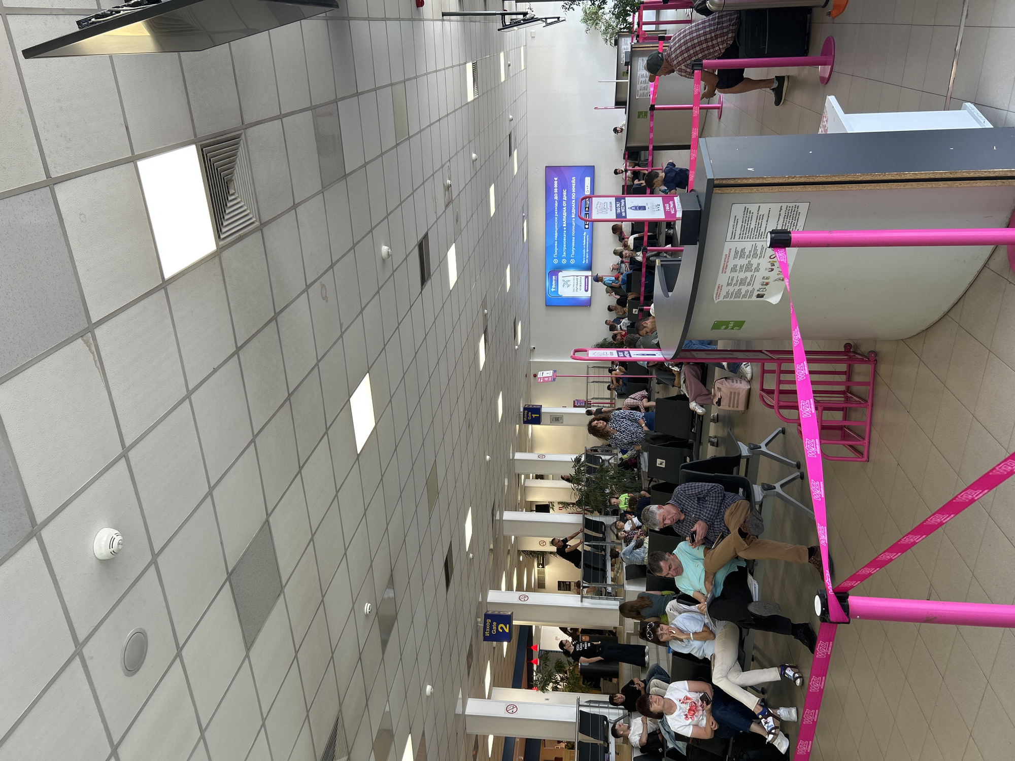 This is a vertical, eye-level shot of an airport waiting area. The scene is dominated by a long, brightly lit corridor with a tiled ceiling and floor. Rows of chairs are set up along one side of the corridor, occupied by a diverse group of people of various ages, some appearing relaxed or bored while waiting.
The dominant color scheme is neutral with stark white ceiling tiles, grey floors, and the occasional pop of color from the pink stanchions and ribbons that delineate queuing areas. A large digital screen displays information in Bulgarian and English, promoting travel insurance. Signs indicate "Изход Gate 2", suggesting this is a departure lounge.
People are casually dressed, some looking at their phones, others conversing or resting. Luggage is visible near some of the passengers. The overall atmosphere is typical of an airport departure lounge, with a sense of anticipation or waiting. The lighting suggests it's daytime, and there are no clear indicators of weather. The presence of the Bulgarian text and the "Sofia, Bulgaria" context place the location firmly in the Sofia airport. A man in a plaid shirt and shorts is standing near a check-in counter. The pink ribbons are branded with "WIZZ", indicating this might be a Wizz Air boarding area.