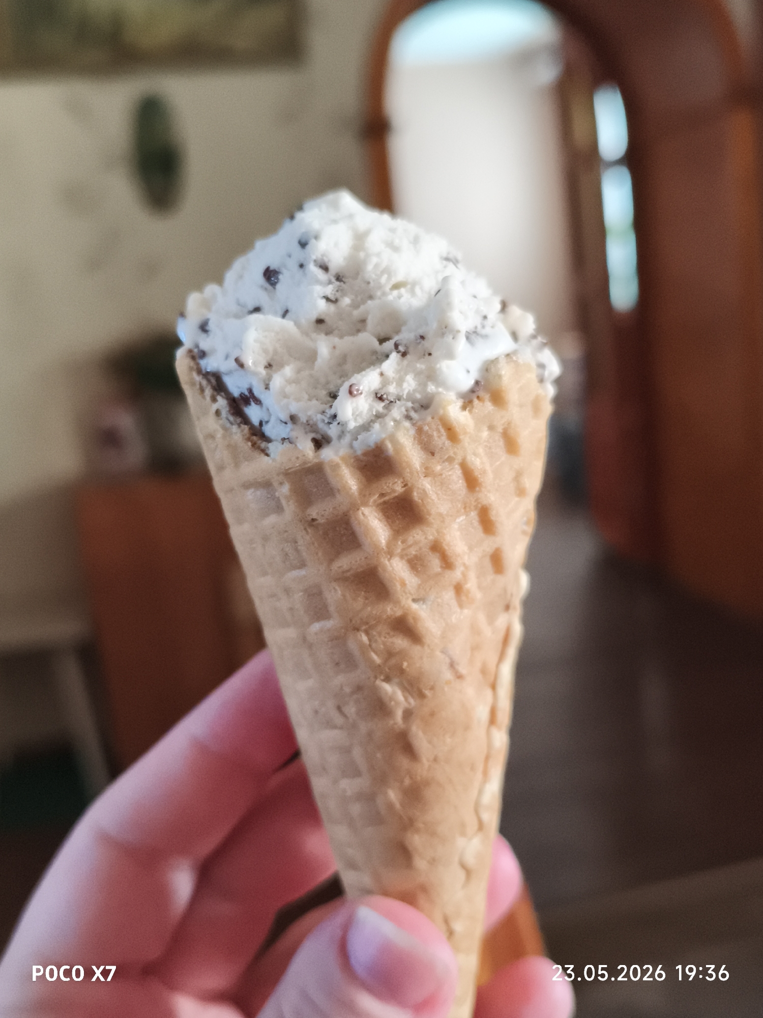 A person holds an ice cream cone in their hand. The ice cream is white with dark chocolate flecks. The cone is a light brown color. The background is blurry, but it appears to be inside a home with a door and some furniture visible. The image also contains the text "POCO X7" in the bottom left corner and the date and time "23.05.2026 19:36" in the bottom right corner. There is no specific indication that this image was taken in the city of Chaikovsky, Russia.