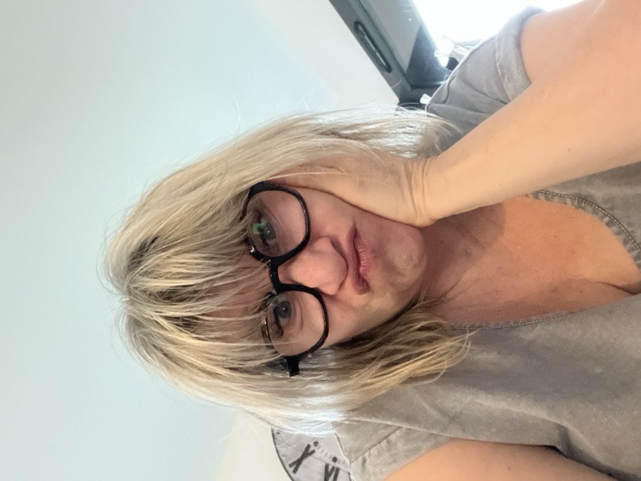 The image displays a close-up, rotated portrait of a person with blonde hair and dark-rimmed glasses. The image is oriented such that the person's head occupies the left side of the frame, with their body extending towards the right.
When mentally rotated upright, the person appears to be a woman, looking upwards towards the camera from a low angle, creating a perspective from below her chin. Her medium-length blonde hair is styled with bangs that partially cover her forehead and frame her face; some darker roots are visible. She wears large, round, dark-framed eyeglasses, through which her eyes are visible. Bright reflections, possibly from an overhead light source or a window, are prominent in the lenses of her glasses. Her mouth is slightly pursed, forming a pout, which suggests an expression of mild annoyance, contemplation, or boredom. Her left hand is pressed against her left cheek and jawline, gently pushing up the skin. She is wearing a casual, short-sleeved grey top, possibly a t-shirt or a chambray shirt, with a visible seam detail near the shoulder or neckline.
The background is largely a light, plain color, likely a wall or ceiling. In the lower right corner of the upright image, a partial view of a large clock face with Roman numerals (specifically 'X', 'XI', and 'XII') is visible. In the upper left background, a dark, rectangular object, which could be part of a screen, a furniture edge, or a window frame, is partially seen, reflecting some light. The overall impression is that of a selfie, taken from a low vantage point, capturing a moment of a casual or contemplative expression.