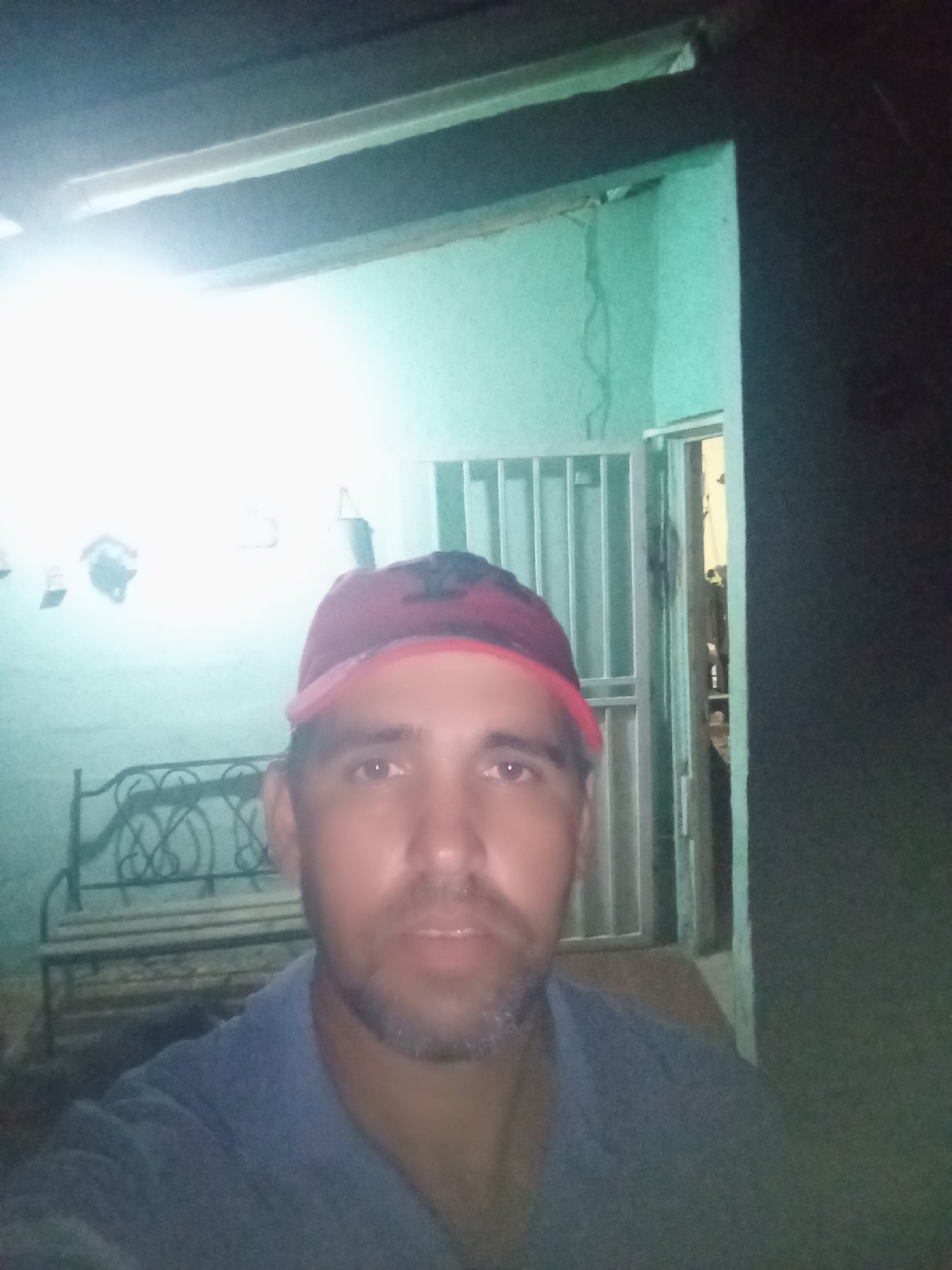 A medium shot depicts a man with fair skin, wearing a red and black baseball cap and a gray t-shirt, positioned indoors. The background shows an interior space with light green walls and a brighter area with a bench and some indistinct objects. A gated doorway is visible behind the man, slightly ajar, hinting at a possible exterior. The scene's dim lighting suggests an evening setting. While the exact location in Juan Griego, Venezuela, is not discernible from the image alone, the architectural style and lighting could be consistent with residential areas in the region.