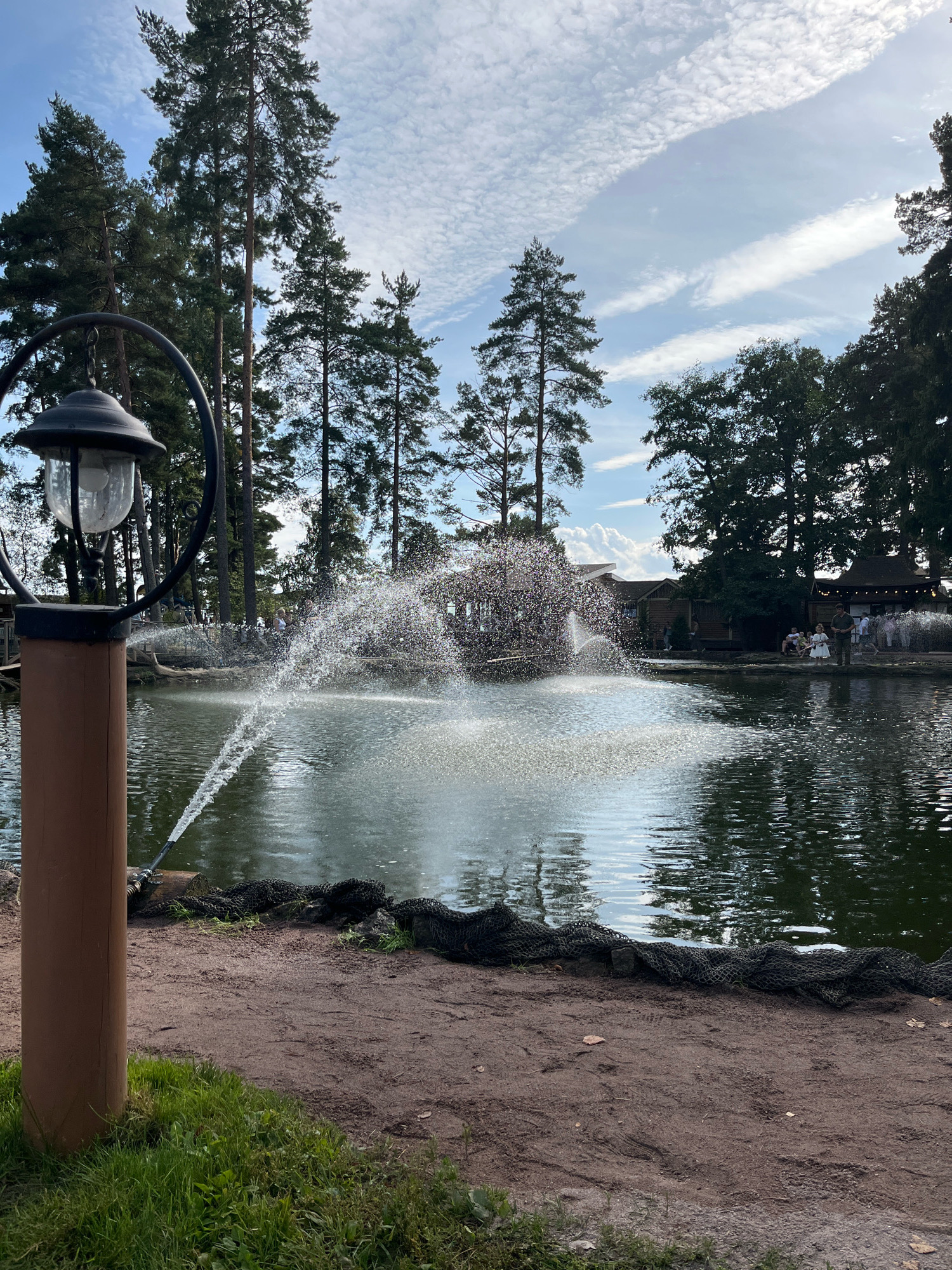 The image depicts a pond with a fountain in a park-like setting in Komarovo, Russia. The fountain sprays water upwards in a wide arc, creating a shimmering effect. To the left of the pond, a tall, dark lamp post stands in the foreground, its glass lantern visible. The pond is surrounded by lush green trees, primarily tall pines, which create a natural backdrop. In the distance, several wooden buildings with thatched roofs are visible, suggesting a recreational area or restaurant. A few people are scattered around the edge of the pond, some sitting and others standing. The sky above is a mix of blue with scattered white clouds. The ground around the pond is a sandy path with some grass and black netting laid along the edge.