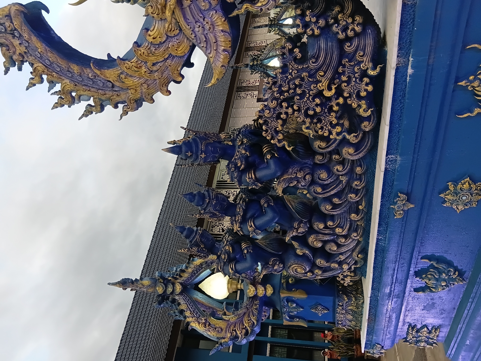 The image depicts a vibrant blue and gold ornate sculpture in front of a building with a dark tiled roof. The sculpture features several figures, possibly mythical beings, with elaborate headdresses and ornate clothing, rendered in a deep blue hue with gold accents. The surrounding architectural elements of the sculpture are also adorned with intricate golden patterns. In the background, the building appears to be a structure with a facade featuring horizontal lines and patterned screens. The sky above is overcast, suggesting a cloudy day. To the left of the sculpture, a lamp with a warm glow is visible. The overall scene suggests a religious or cultural site, possibly a temple, given the artistic style and the detail of the sculpture. Based on the style of Buddhist art, it is likely located at Wat Rong Khun (White Temple) or Wat Rong Suea Ten (Blue Temple) in Chiang Rai City Municipality, Thailand.