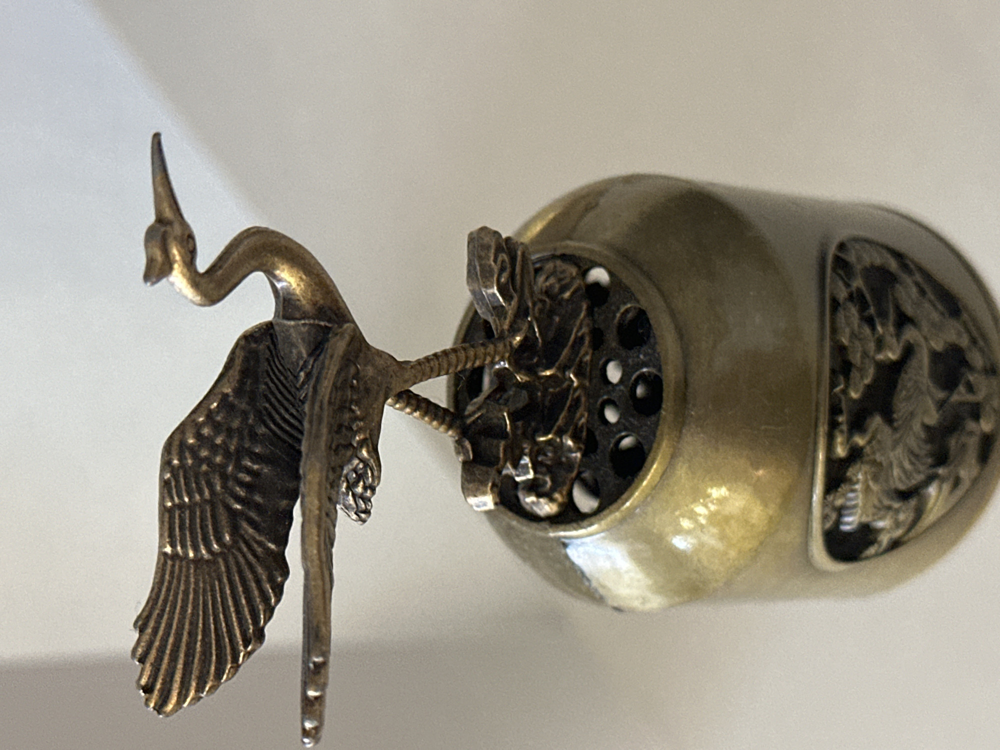 The image shows a close-up of an ornate, antique-looking brass ornament. The ornament is topped with a detailed sculpture of a bird, possibly a crane or heron, with its wings spread and head held high. The bird is perched on a circular base with intricate carvings and multiple small holes, suggesting it might be part of an incense burner or a similar decorative item. The lower portion of the ornament, visible in the image, is cylindrical and features a decorative band with a relief carving of mythical creatures, likely dragons or other serpentine beings. The overall impression is one of craftsmanship and traditional artistry. The background is a plain, light-colored surface, out of focus, which emphasizes the detailed subject. There is no indication of the ornament's location within the city of Nevinnomyssk, Russia, or any context related to its environment.