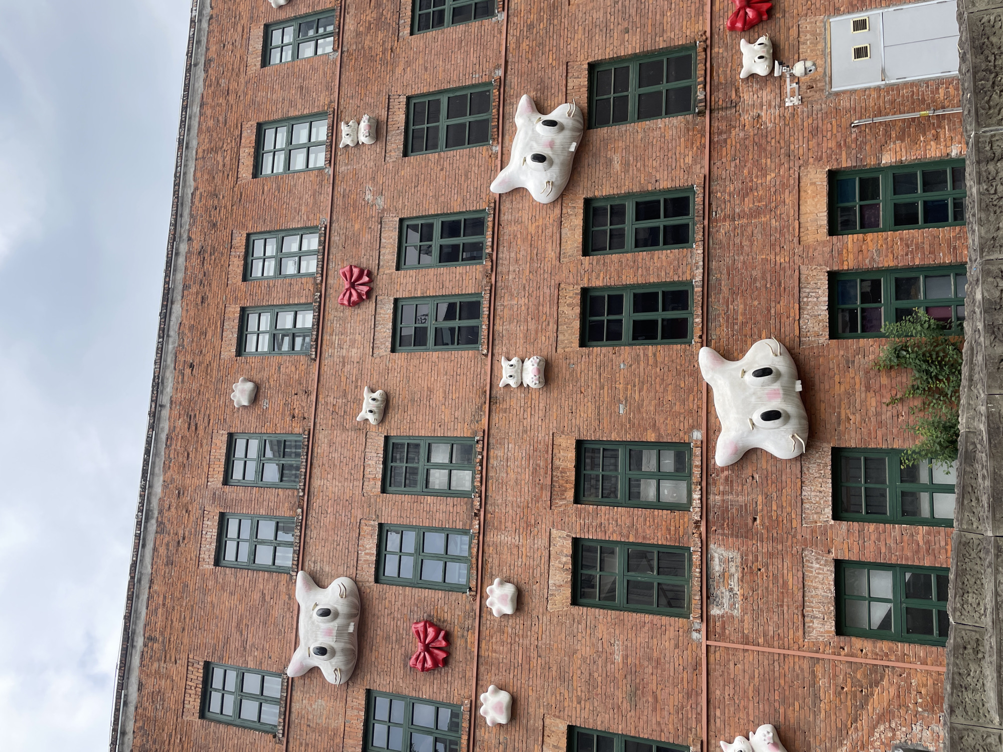 A low-angle, medium shot depicts a red brick building adorned with whimsical sculptures in Yuzhong District, China. The building features multiple rows of green-framed windows. Plaster-like sculptures resembling cartoonish ghosts or creatures with large eyes, smaller abstract figures, and red bow shapes are affixed to the exterior. The sky is overcast, with visible cloud cover.