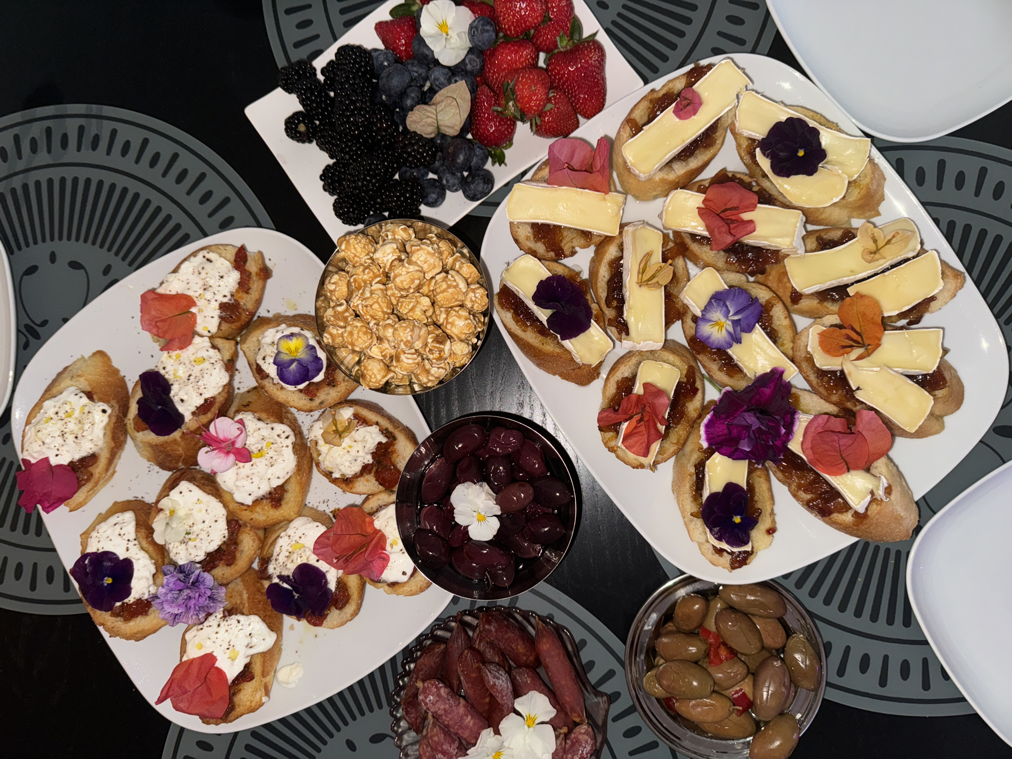 The image shows an overhead view of a variety of food items arranged on a dark surface with circular gray patterned placemats.
Food items include:
* A white rectangular plate containing strawberries, blueberries, and blackberries.
* A glass bowl filled with caramel popcorn.
* A white plate with toasted baguette slices topped with a white, creamy substance and decorated with colorful edible flowers.
* A white plate with toasted baguette slices topped with a brownish spread and Brie cheese, also decorated with edible flowers.
* A stainless steel bowl filled with dark olives, garnished with a white flower.
* A decorative glass plate with small, cured sausages decorated with white flowers.
* A stainless steel bowl filled with green olives and red peppers.
The background is dark. The image does not provide enough information to pinpoint a location in Belgrade, Serbia, or to determine the presence of people, actions, interactions, or further environmental details beyond the food arrangement.