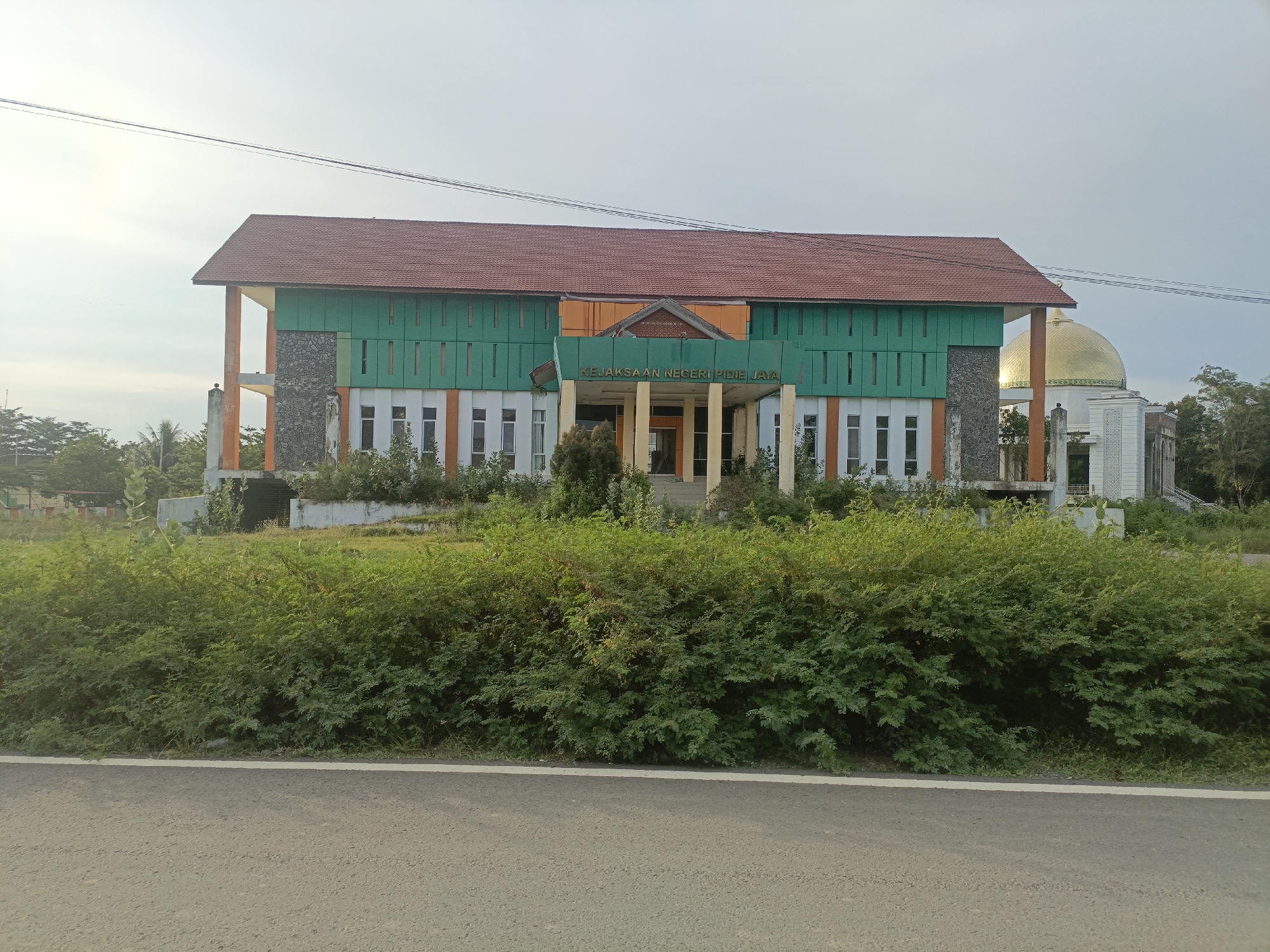 In the city of Meureudu, Indonesia, an outdoor, eye-level medium shot captures a building with a prominent green facade and a red tile roof. The building is situated behind a dense line of green foliage, with a paved road and a white line running along the bottom of the frame. To the right of the main building, a golden dome, likely belonging to a mosque, is visible. The sky above is overcast with a faint hazy glow. The building appears to be an institutional structure, possibly a government office, as evidenced by the text "KEJAKSAAN NEGERI PIDIE JAYA" visible on the facade. The overall scene suggests a daytime setting under a cloudy sky, with the natural environment of lush greenery contrasting with the man-made structures.