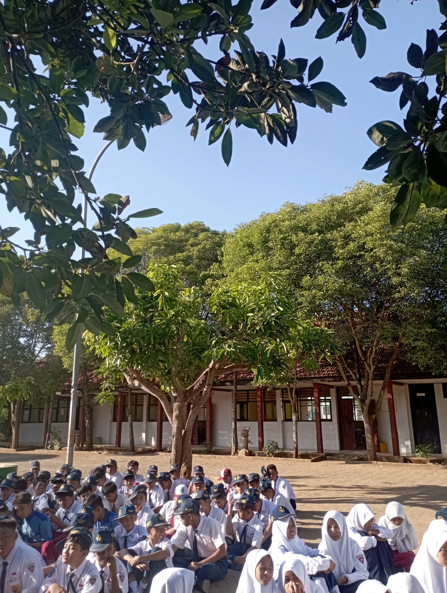 The image shows an outdoor gathering of students, likely in a schoolyard, with trees and buildings in the background. The students, appearing to be of elementary or middle school age, are dressed in uniforms, many with white shirts and dark pants or skirts, and some wearing caps. They are seated on the ground in a relaxed manner, some facing the camera, others looking in different directions. The environment appears to be warm and sunny, with clear blue skies visible through the leafy branches of trees that frame the top of the image. The buildings in the background are low-rise structures with visible windows and roofs, suggesting an educational or institutional setting. The overall atmosphere appears to be one of an informal assembly or break. The location is identified as Banyuurip, Indonesia.