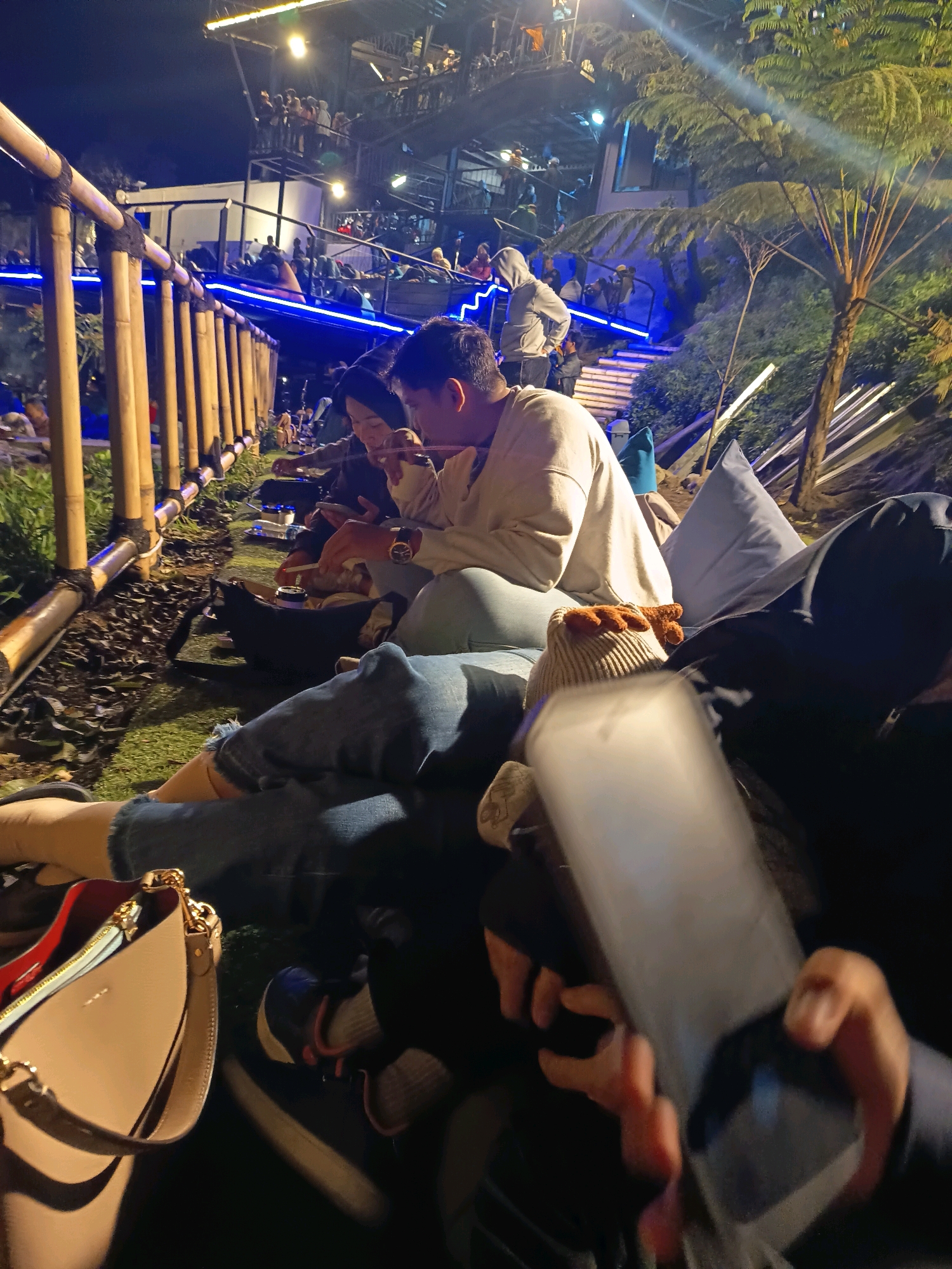 In Keditan, Indonesia, a group of individuals is seen seated outdoors at night, with a multi-level structure and illuminated railings in the background. The foreground features people lounging on the grass, some engaged with their phones, while others are interacting with each other. A beige handbag with a gold chain rests on the ground near a person's legs. A tree with large, fern-like leaves is visible on the right side of the frame. The scene suggests a casual social gathering in an outdoor setting, possibly a restaurant or recreational area.