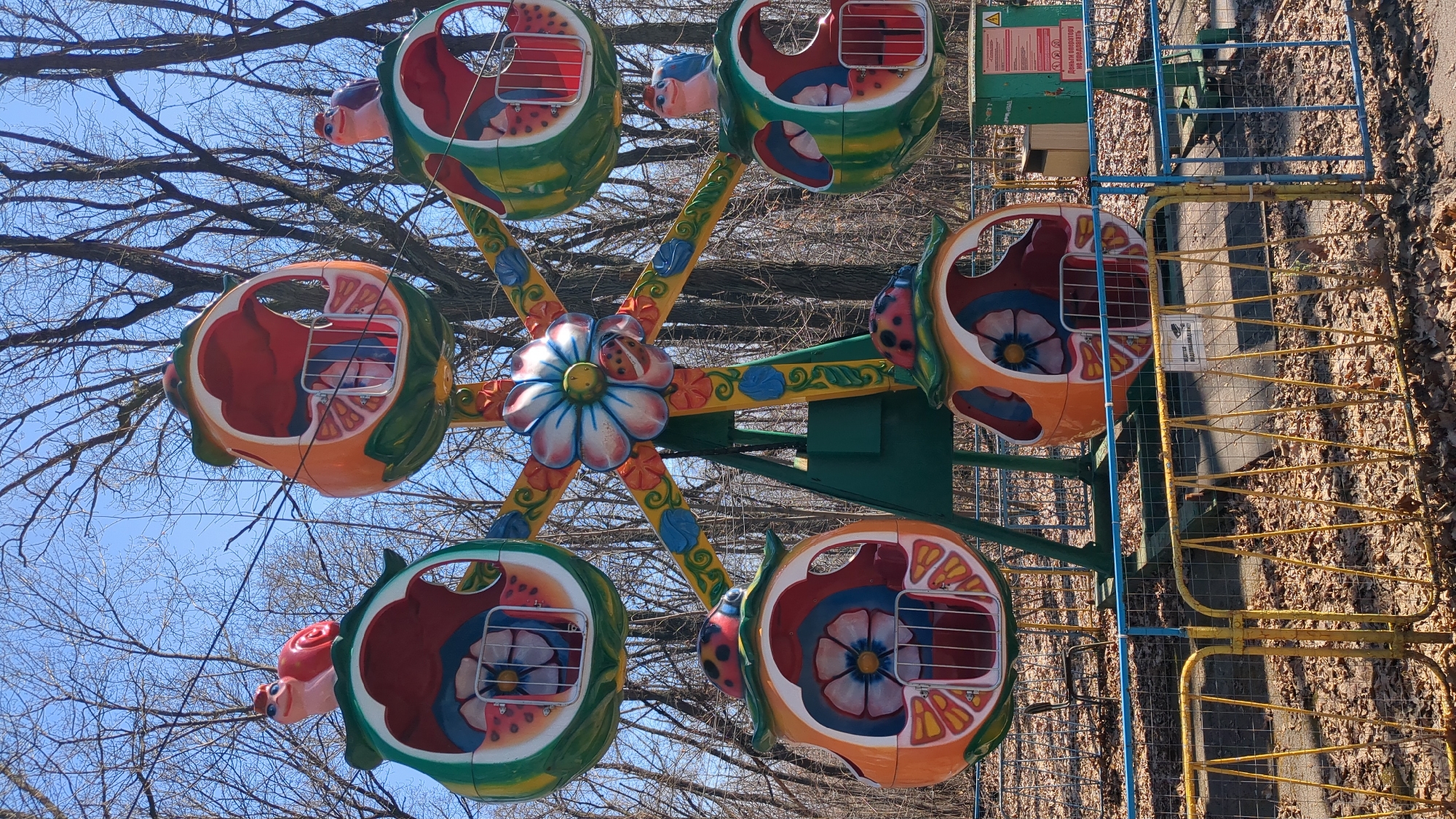 The image shows a colorful, small Ferris wheel likely located in a park or amusement area. The wheel has five fruit-shaped carriages, each with a cartoonish animal or insect figure attached (snails, birds, ladybugs). The carriages are painted in vibrant colors like orange, red, green, and blue. The supporting structure is painted green with decorative floral patterns in orange and blue. The overall impression is that it is designed for young children. The surrounding environment consists of bare trees, suggesting it is either late autumn, winter, or early spring. There is a fenced area nearby made of rusty yellow metal. The location could be the Park named after Vladimir Nikolaevich Tretjakov, in the city of Ulyanovsk, Russia.