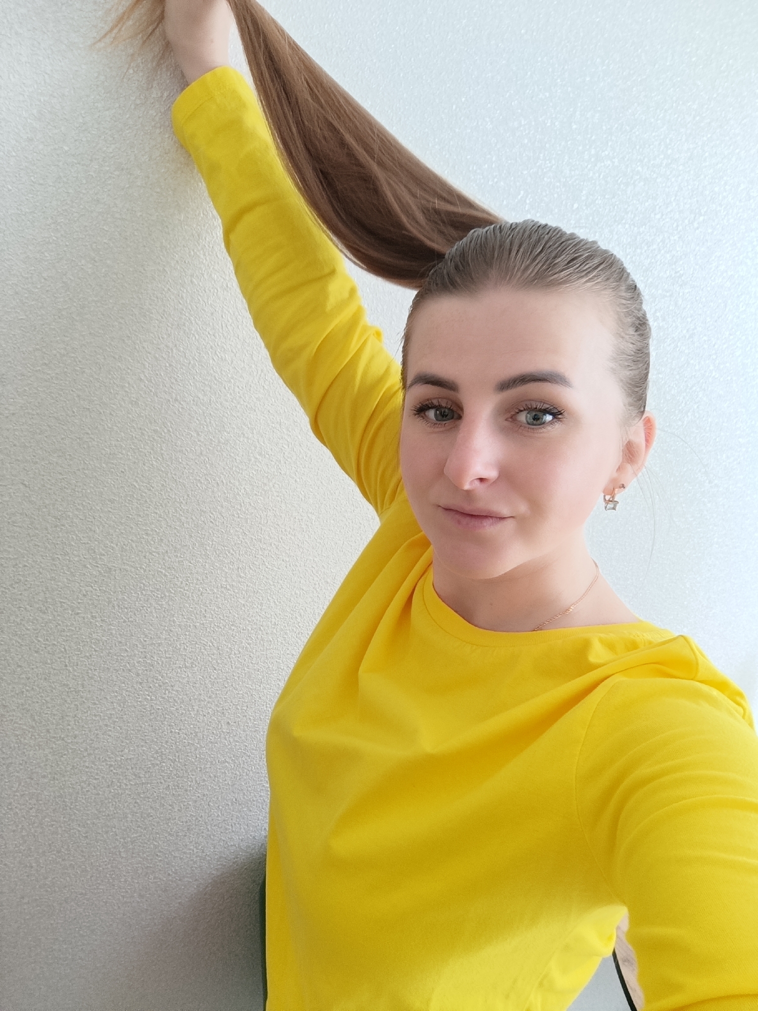 A young woman stands indoors against a light-colored, textured wall. She is wearing a bright yellow long-sleeved shirt and has her brown hair pulled back in a high ponytail, which she is holding aloft with her right hand. She is looking directly at the camera with a slight smile. Her face is clearly visible, showing green eyes and light skin. She is wearing earrings and a delicate necklace. No specific details indicate the exact location is Chernogolovka, Russia.