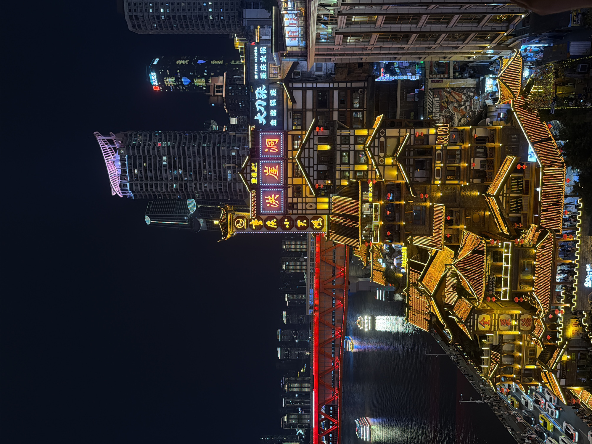 The image captures a nighttime panoramic view of the Hongyadong folk custom area in Yuzhong District, Chongqing, China. The scene is dominated by the illuminated architecture of Hongyadong, characterized by its multi-story, traditional Chinese-style buildings with tiered roofs, adorned with countless warm yellow lights. These structures are built on the side of a hill, overlooking a wide river, likely the Yangtze or Jialing River.
Across the river, a prominent red illuminated bridge spans the water, with vehicles visible crossing it. Several boats are on the river, some illuminated and reflecting on the water's surface. In the background, modern skyscrapers rise into the dark night sky, their windows lit, forming a stark contrast with the historic architecture of Hongyadong.
The foreground shows a busy street with cars and pedestrians. The overall atmosphere is one of vibrant urban nightlife, with the interplay of traditional and modern architectural styles, illuminated by a multitude of lights. The scene is depicted from an elevated vantage point, offering a sweeping perspective of the bustling cityscape.